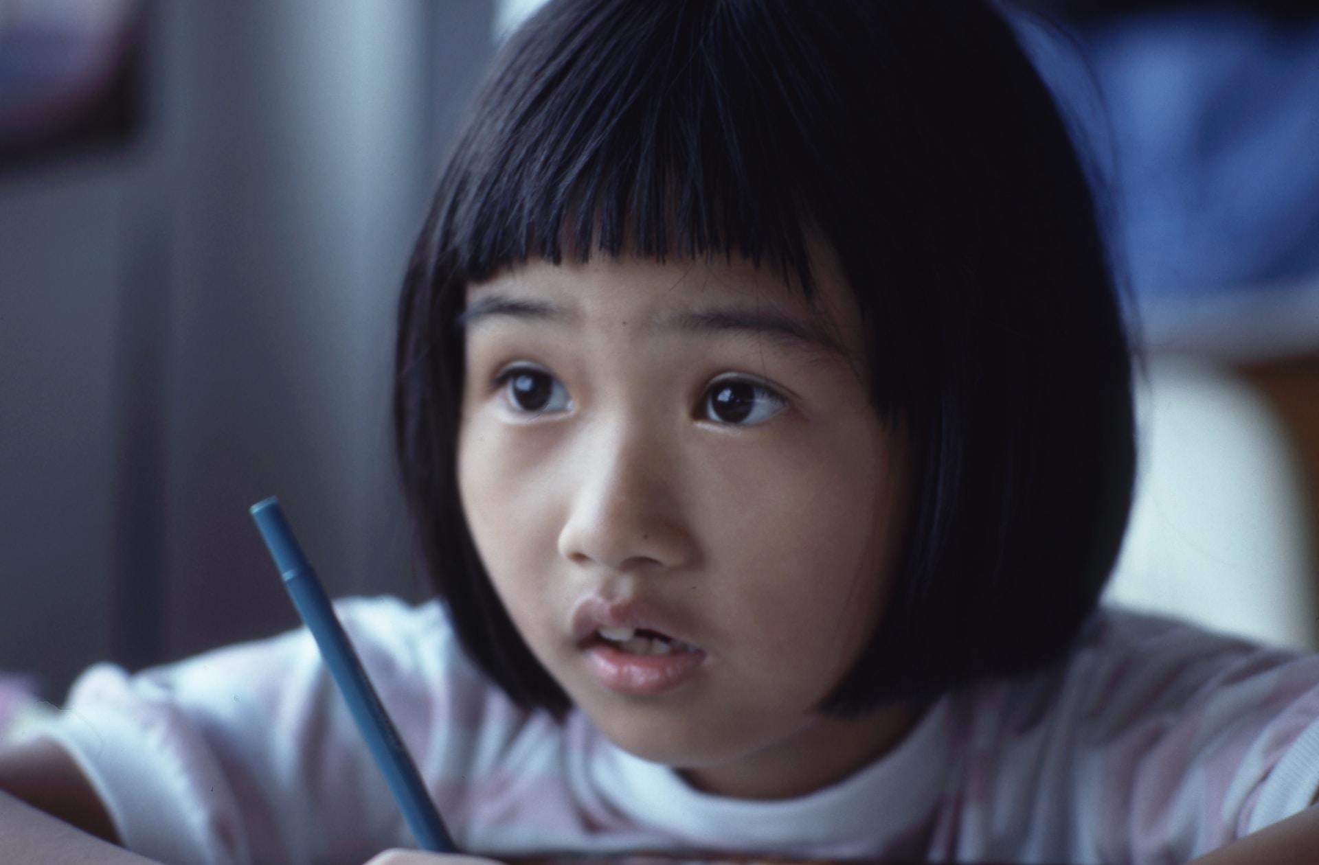A child wearing a pink and white striped top holds a blue pen in their hand while looking up, mouth slightly agape.