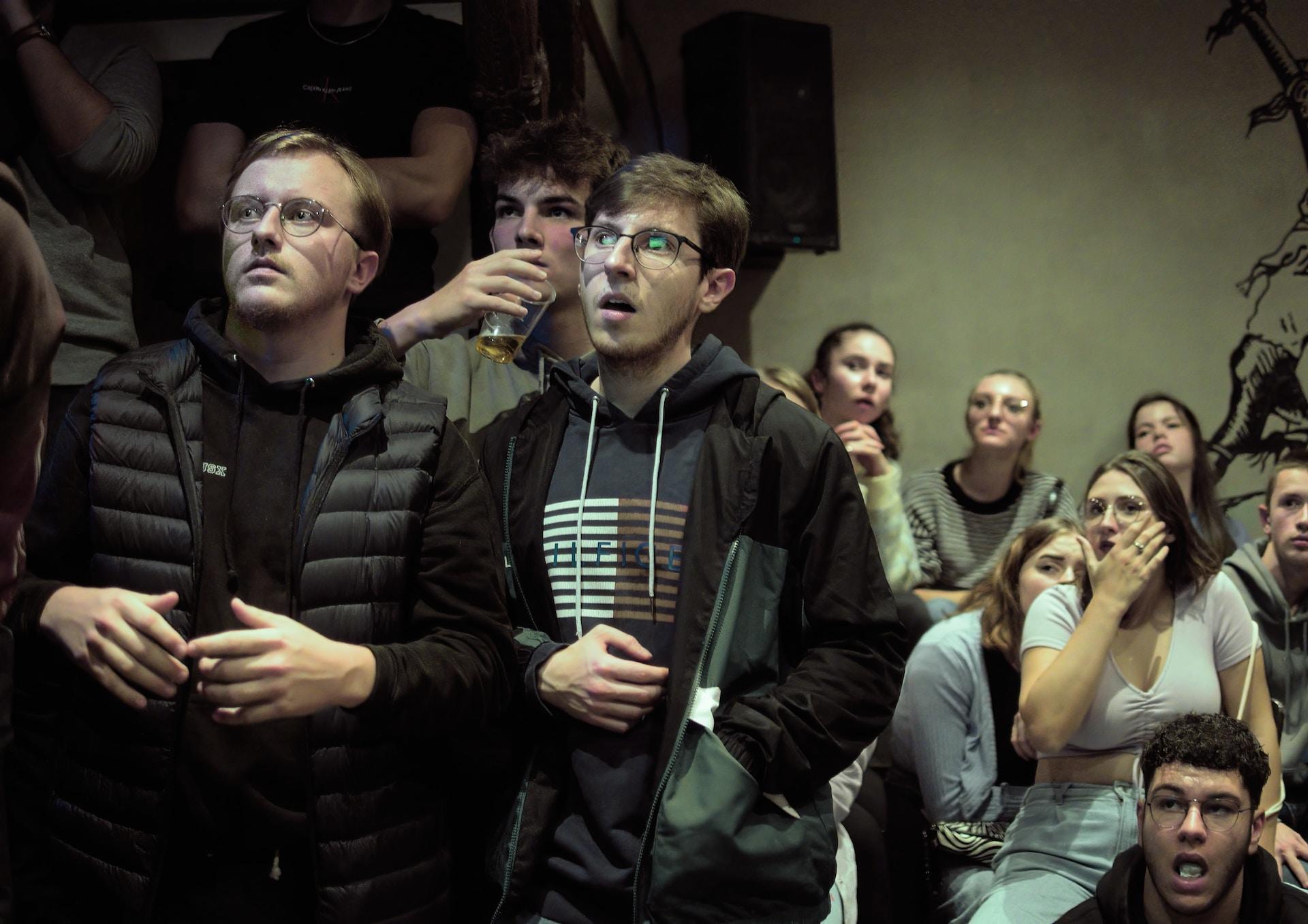 A group of soccer fans in a drab brown room, all wearing muted colours, watching a match with a look of shock on their faces. 