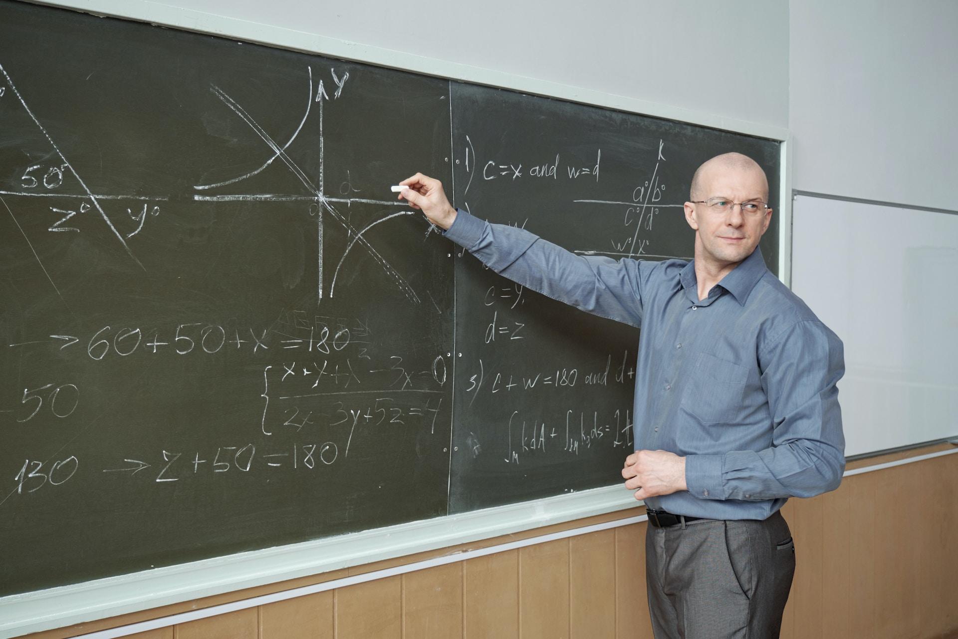 A person in a blue shirt points at a chalkboard with equations written on it with their right hand as they look over their left shoulder.