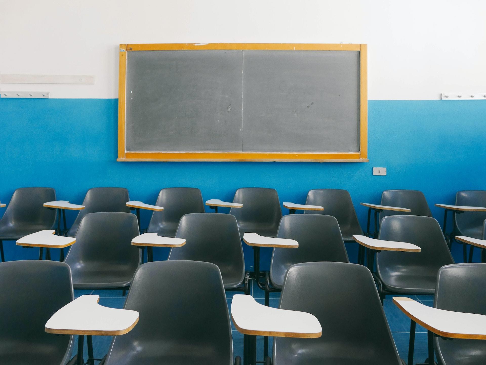 An old-style classroom with bright blue and white walls and a chalkboard hanging behind rows of empty blue chairs, each with a desk attached. 