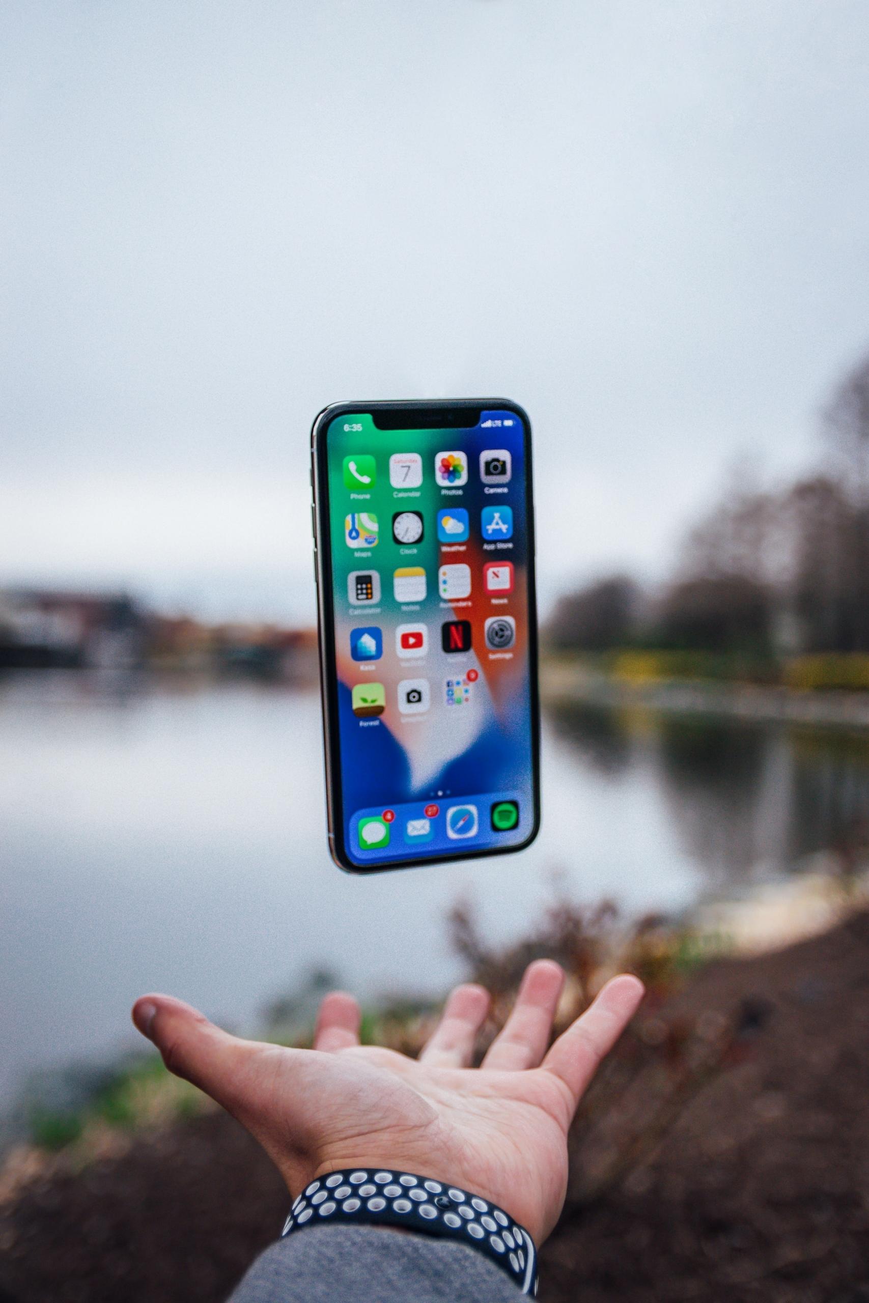 A smartphone with its screen active and displaying numerous icons floats over its owner's outstretched hand against a lakeside background.