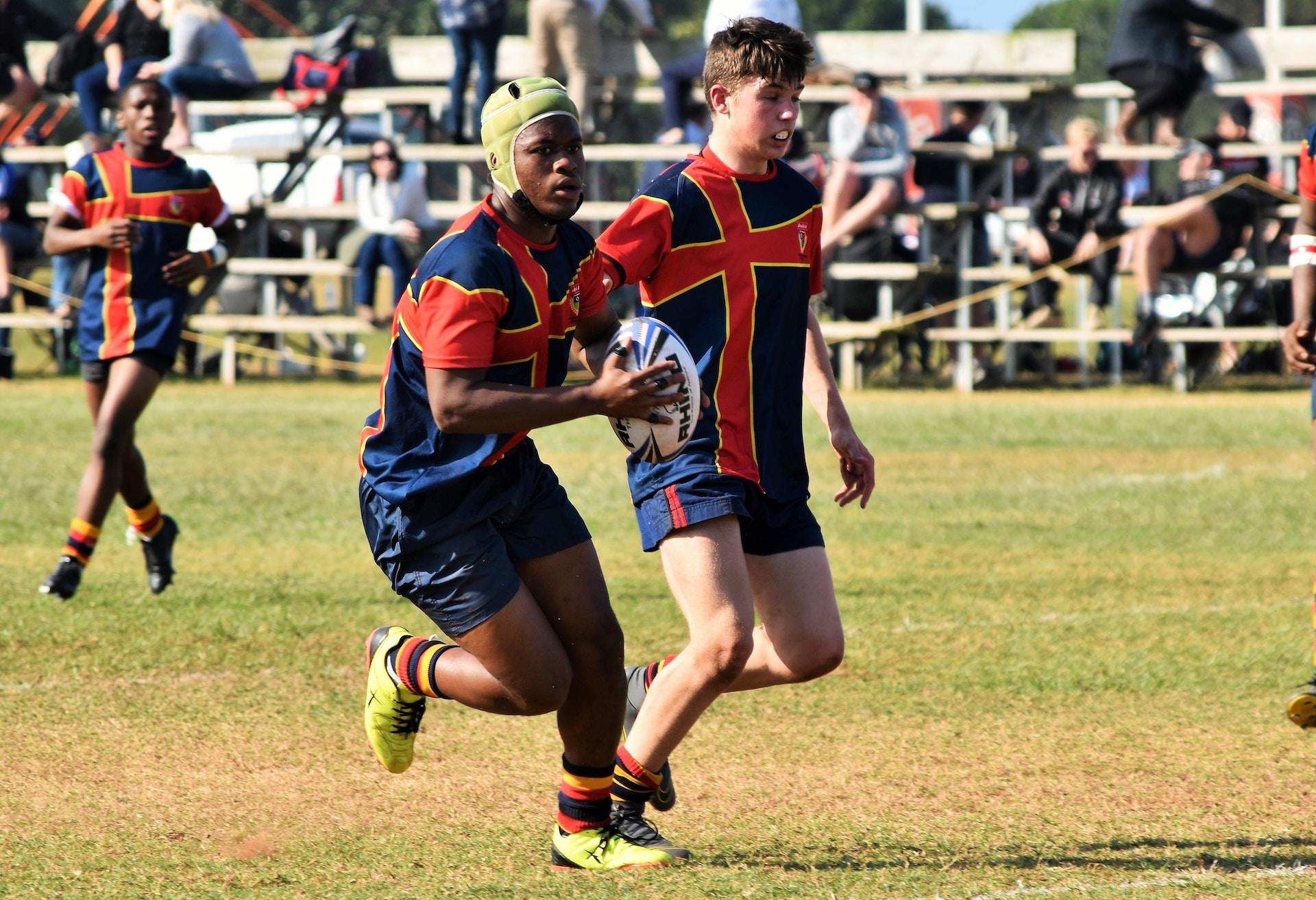 Two rugby players wearing red and blue team uniforms race downfield with the ball. One of them wears a yellow cap. 