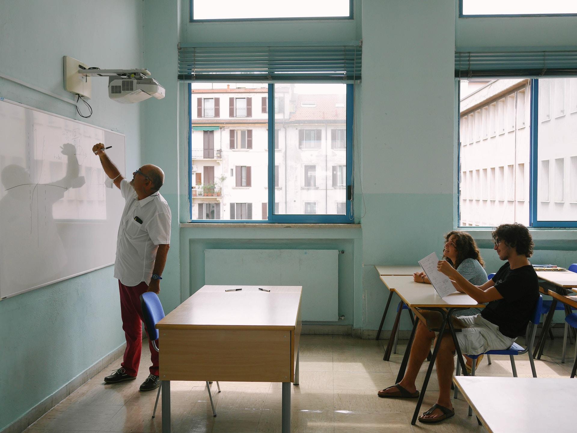 A bare-bones classroom with light-coloured school furniture and institutional green walls, with a person wearing a white shirt writing on the dry-erase board as two people, seated at student desks, look on.