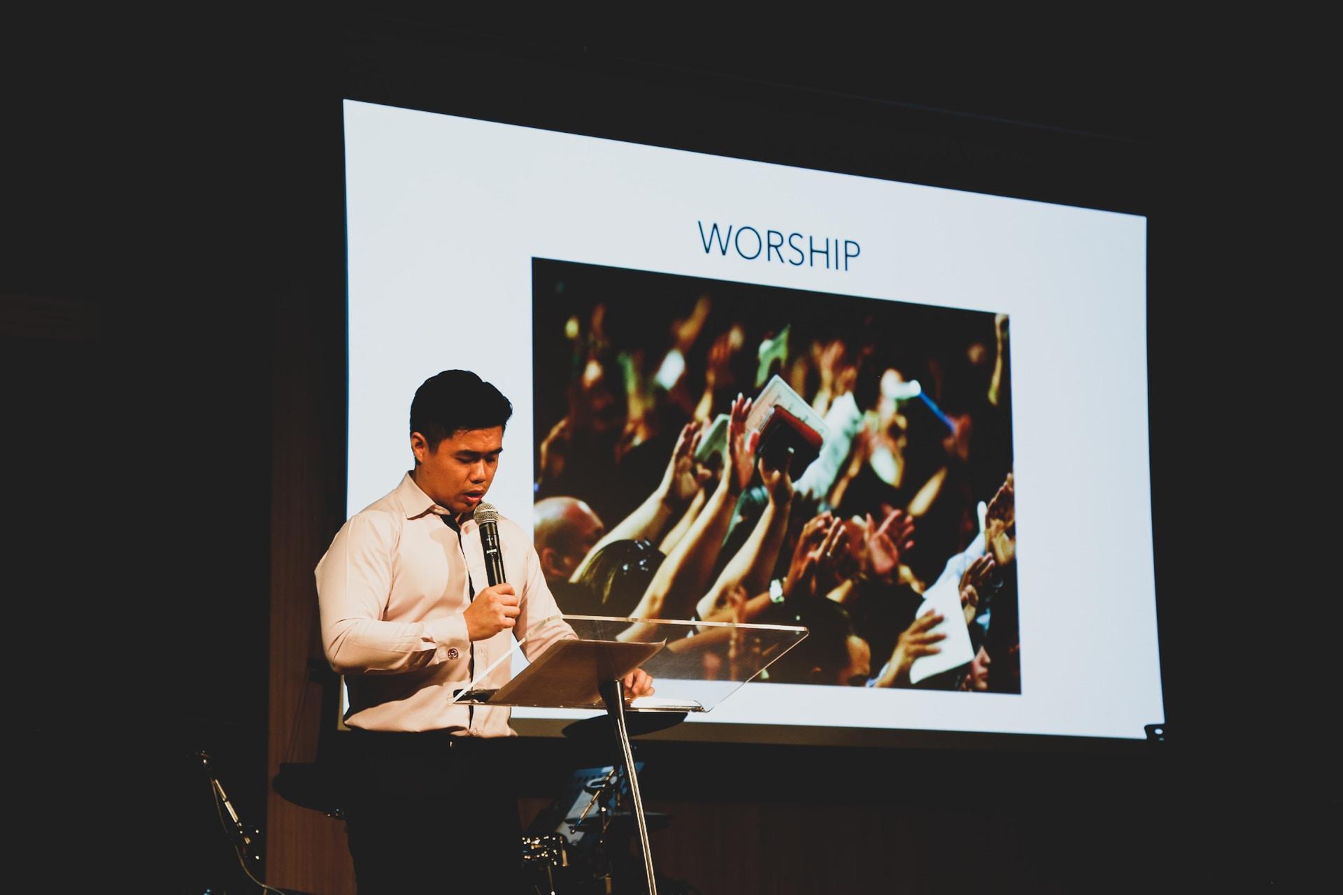 A person wearing a white shirt stands at a lectern in front of a slideshow that says worship.