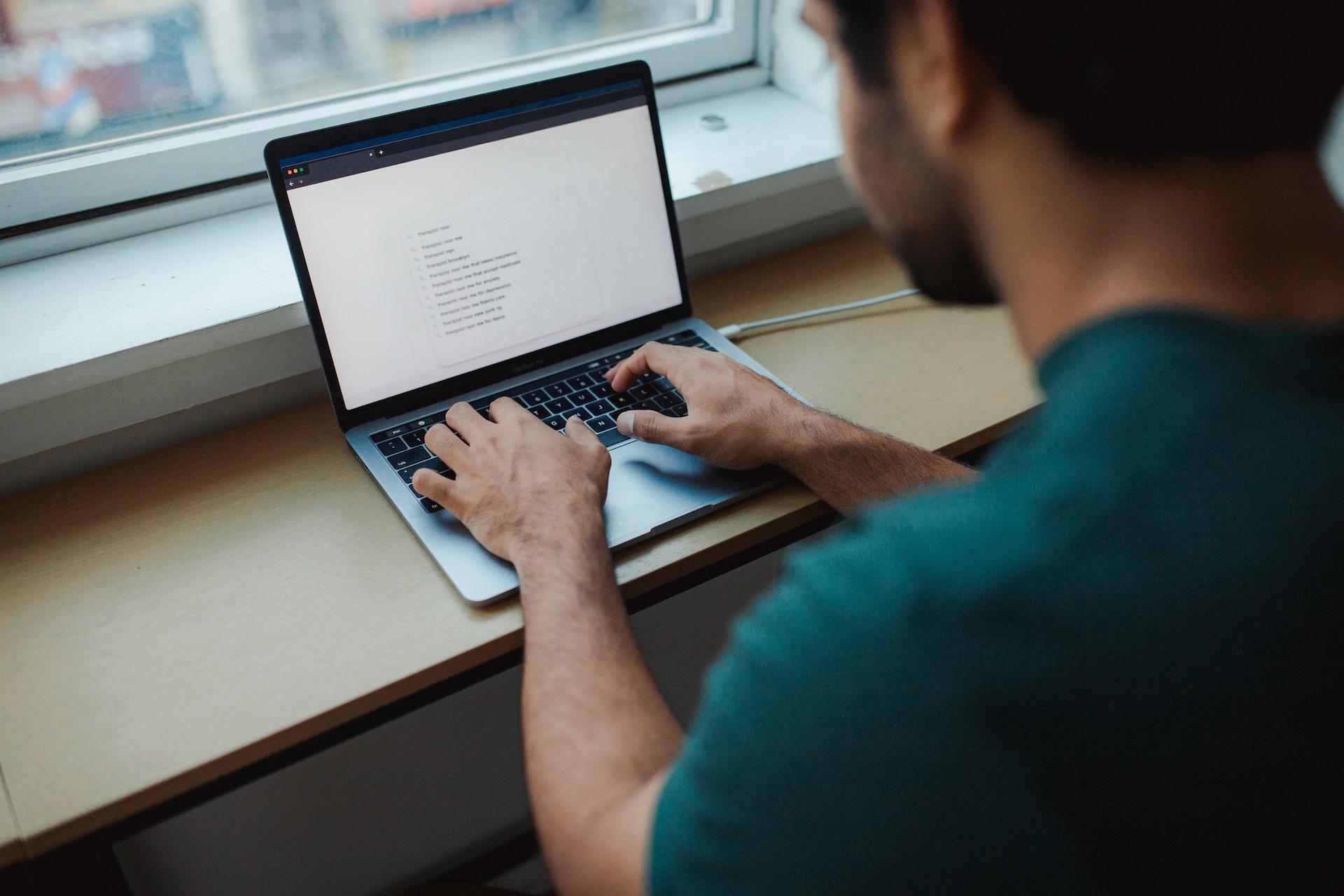 A person in a green short-sleeved top sits at a window with an open laptop poised on a light-coloured ledge, preparing to type a search string into the query box.