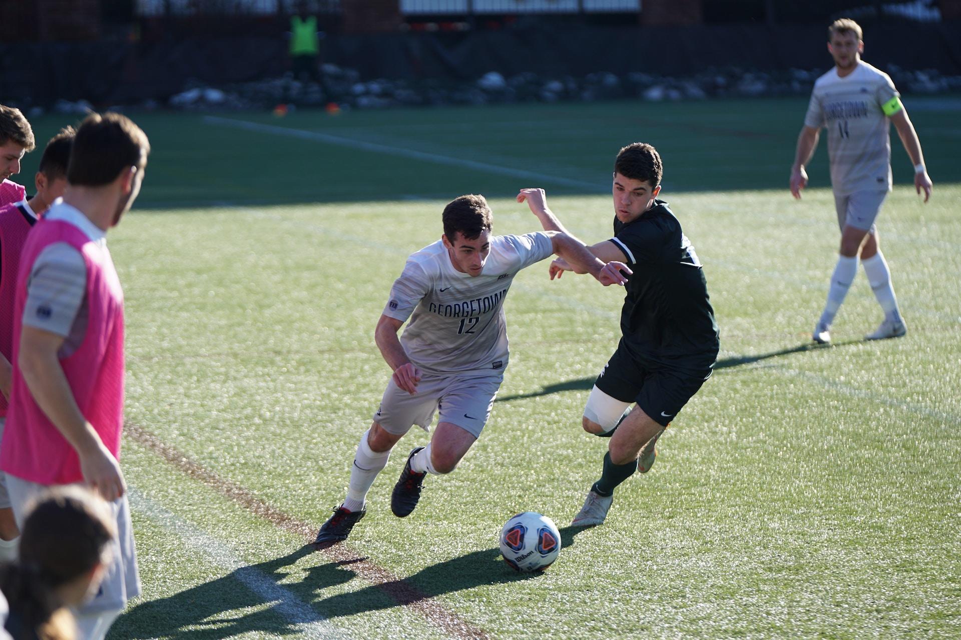 Two soccer players vie for the ball on the green pitch on a sunny day while a pink-shirted captain and a grey-uniformed monitor look on.