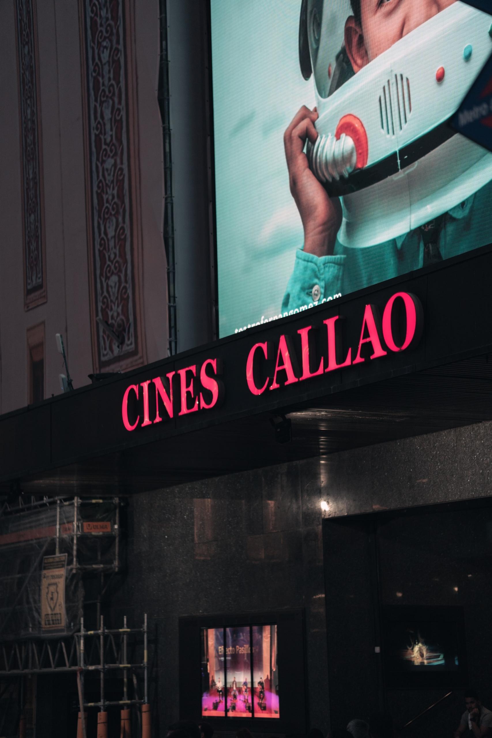 A Spanish movie theatre in nighttime Madrid with its name lit up in pink neon, which contrasts with the turquoise marquee above.