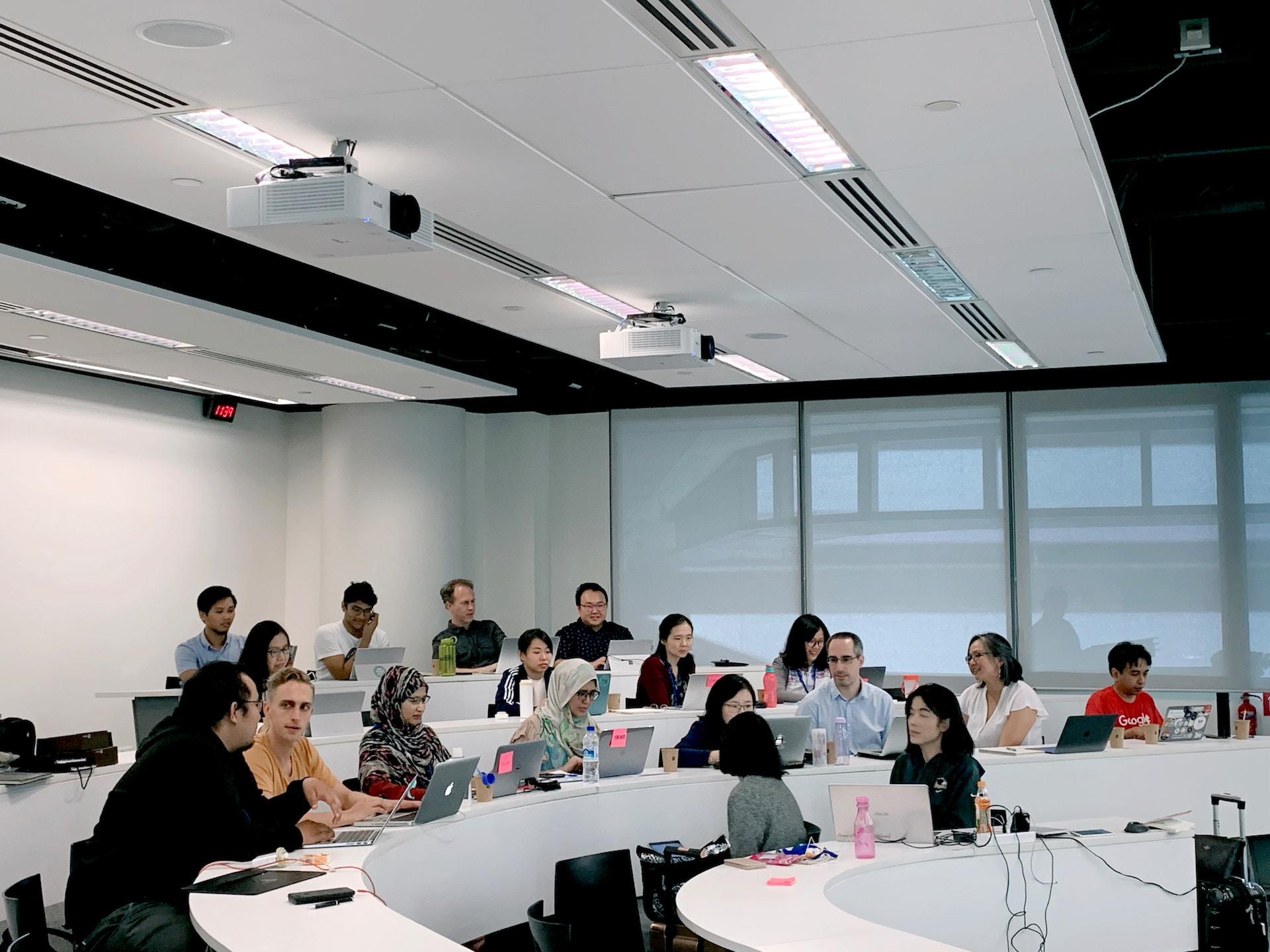A modern-looking classroom with white bench desks curving around a front of the room well and students sitting in a random arrangement.