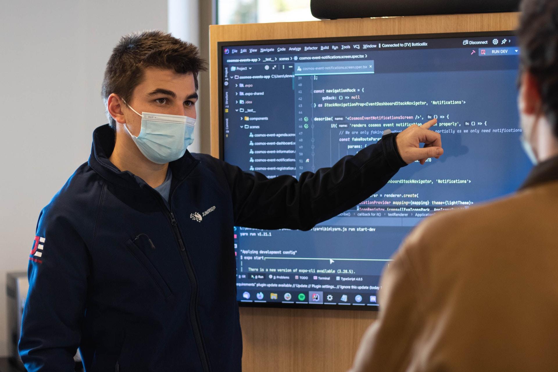 A student wearing a dark coloured top and a face mask stands next to a plasma display showing lines of code. they're pointing at a line of code with their left hand while a person in a brown top looks on. 