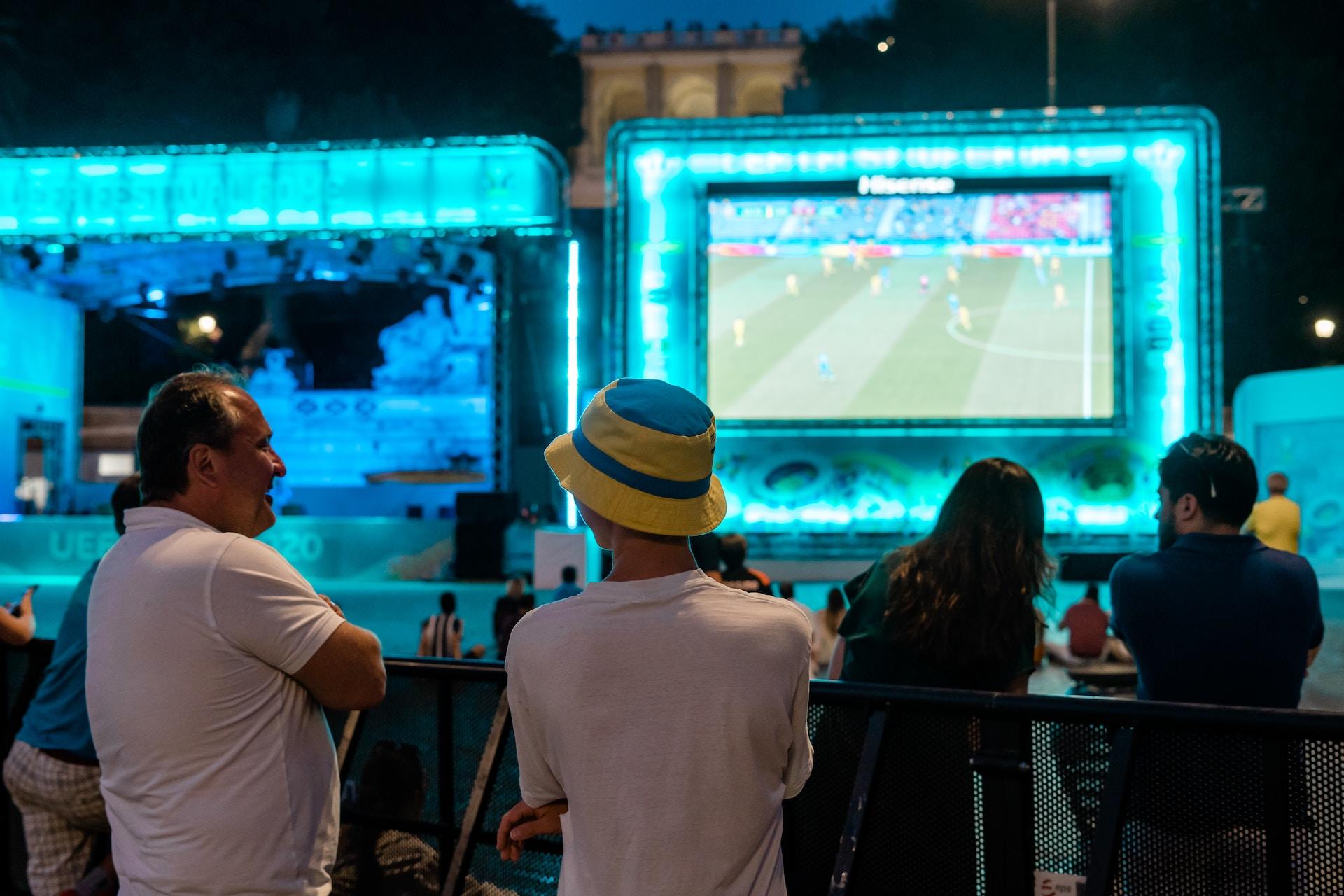 A group of soccer fans gather loosely outside of a venue to watch a soccer match on a screen surrounded by a bright, neon blue frame at night. 