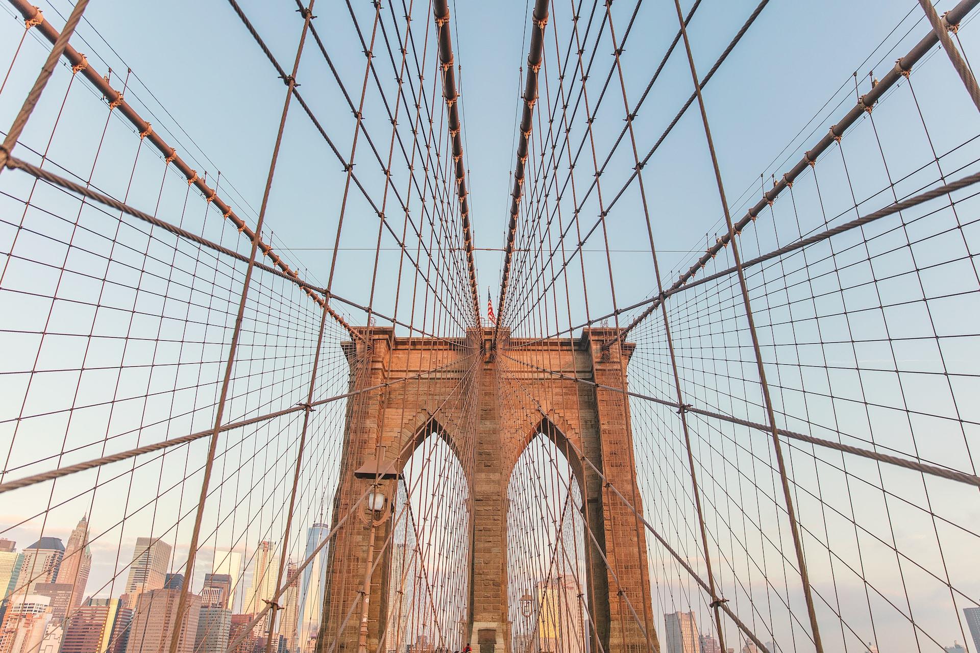 A dual-arch red stone structure forms the joining point for a suspension bridge's cables, which are seen contrasted against the blue sky, with a cityscape in the background. 