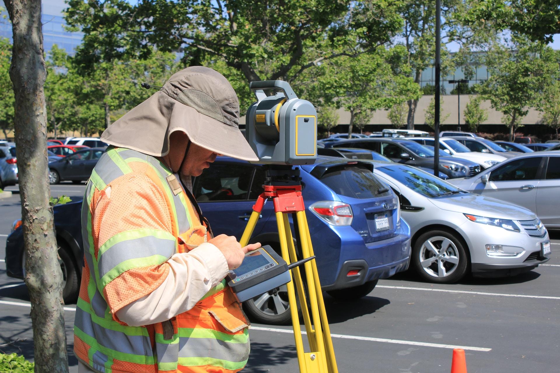 A person wearing a safety vest and a floppy brimmed hat stands by a yellow surveying tripod set up in a parking lot, looking down at its portable console they hold in their hands. 