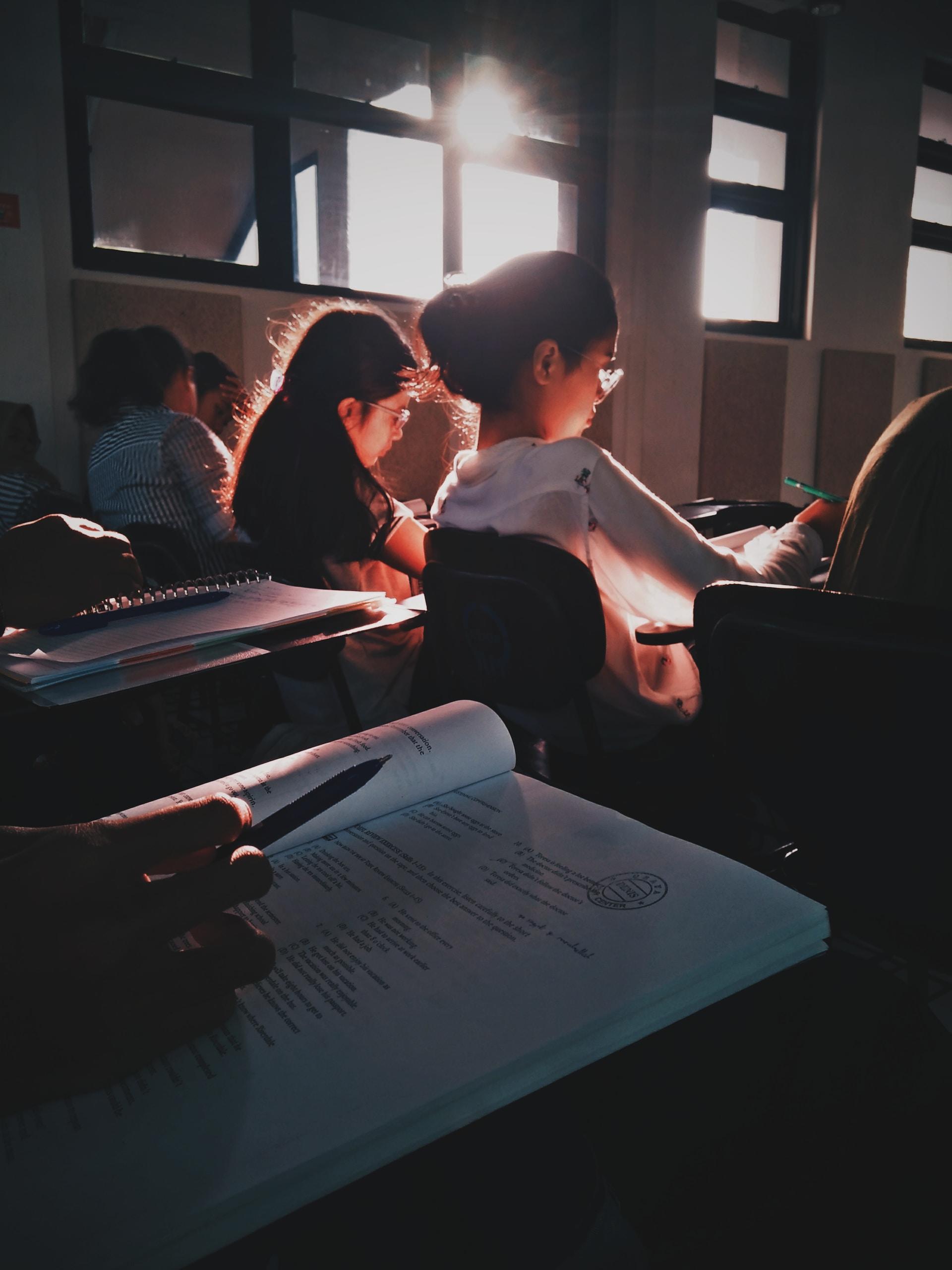 Students sit in a semi-darkened classroom with the sun stabbing the windows. They are focused on their exam papers. 