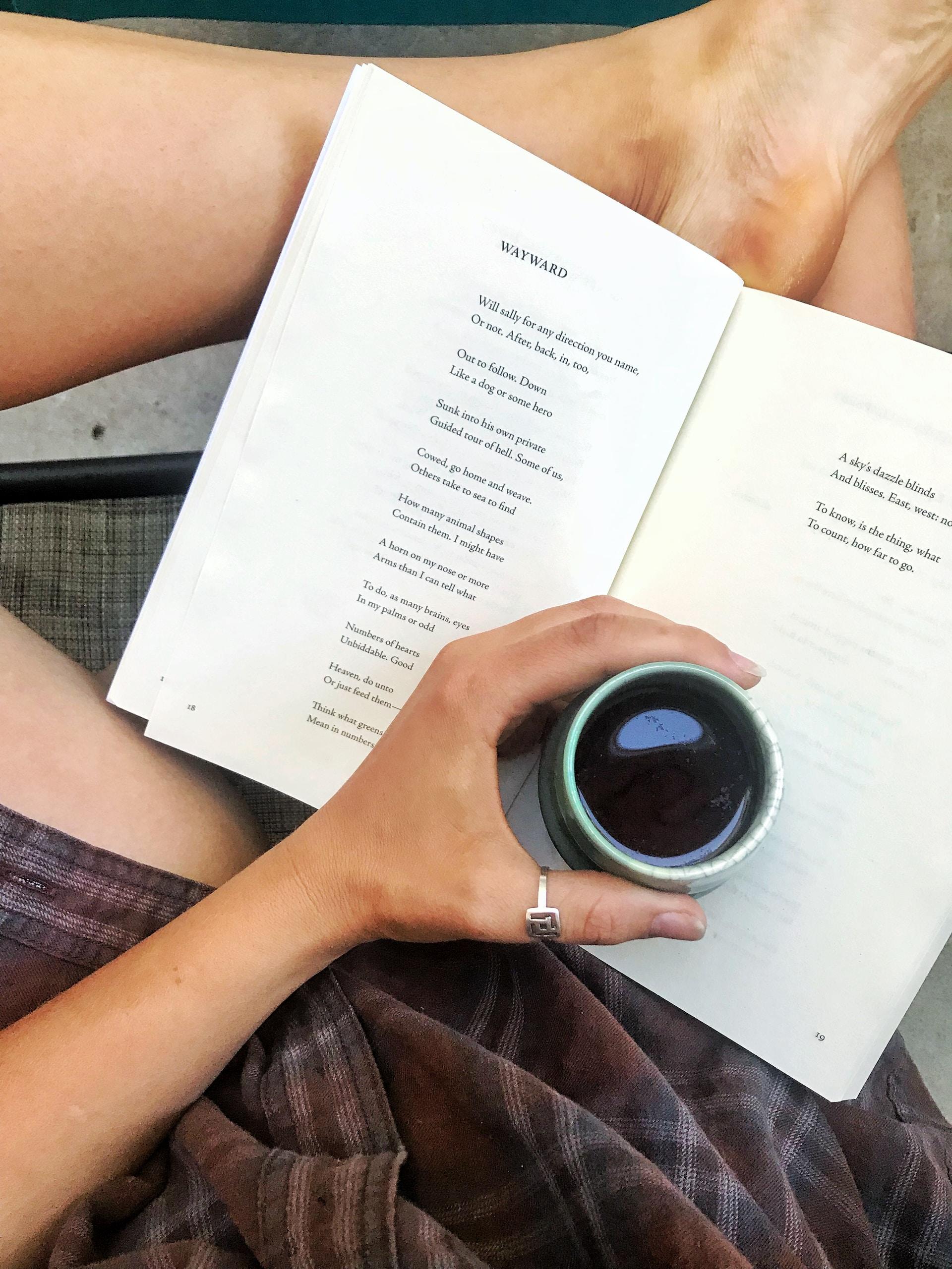 A person wearing a brown plaid garment holds a cup of some liquid over an open poetry book, which is propped up on their legs. 