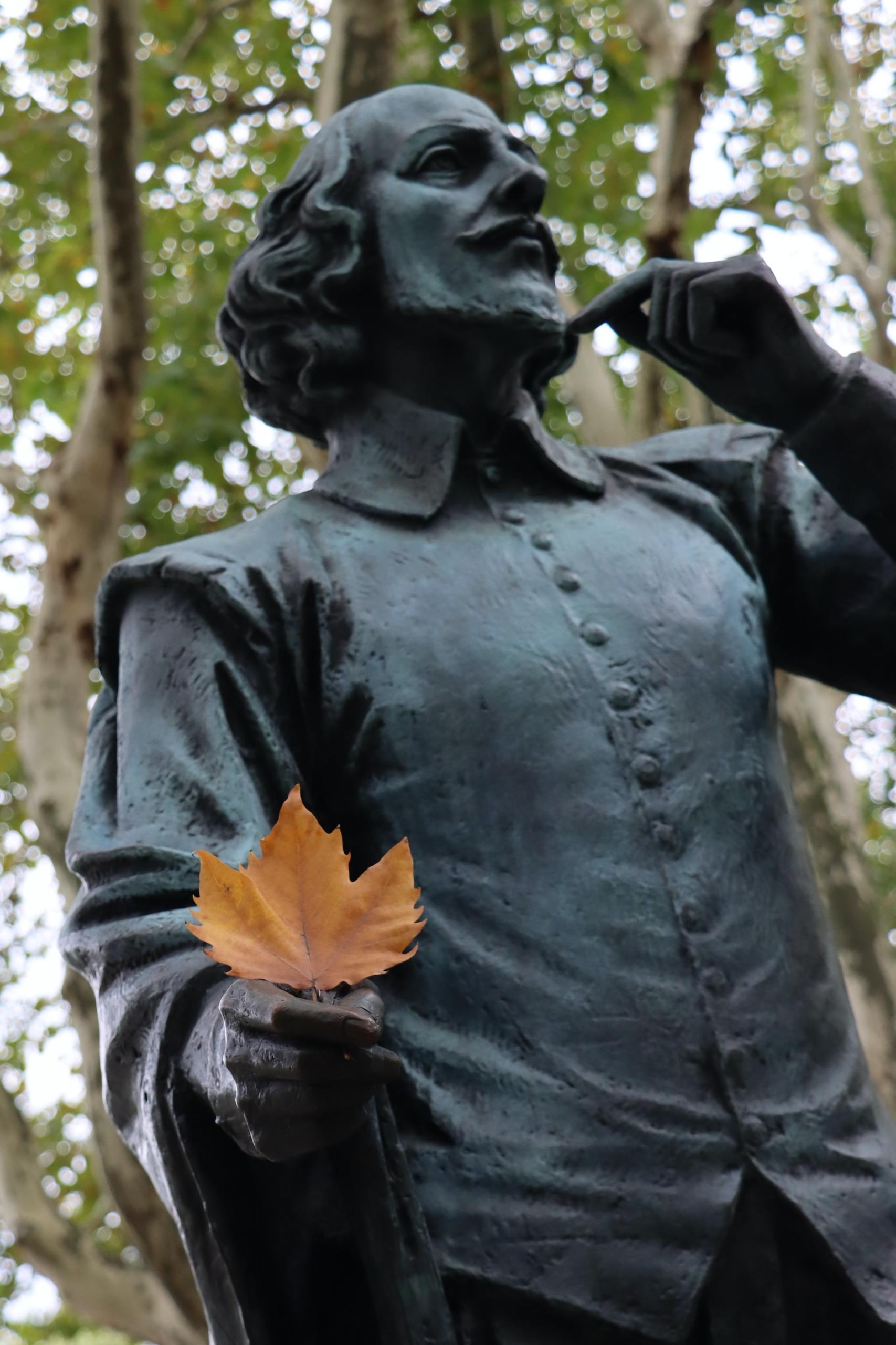 A statue of William Shakespeare with his left index finger pointing to his chin while his right hand holds an orange leaf.