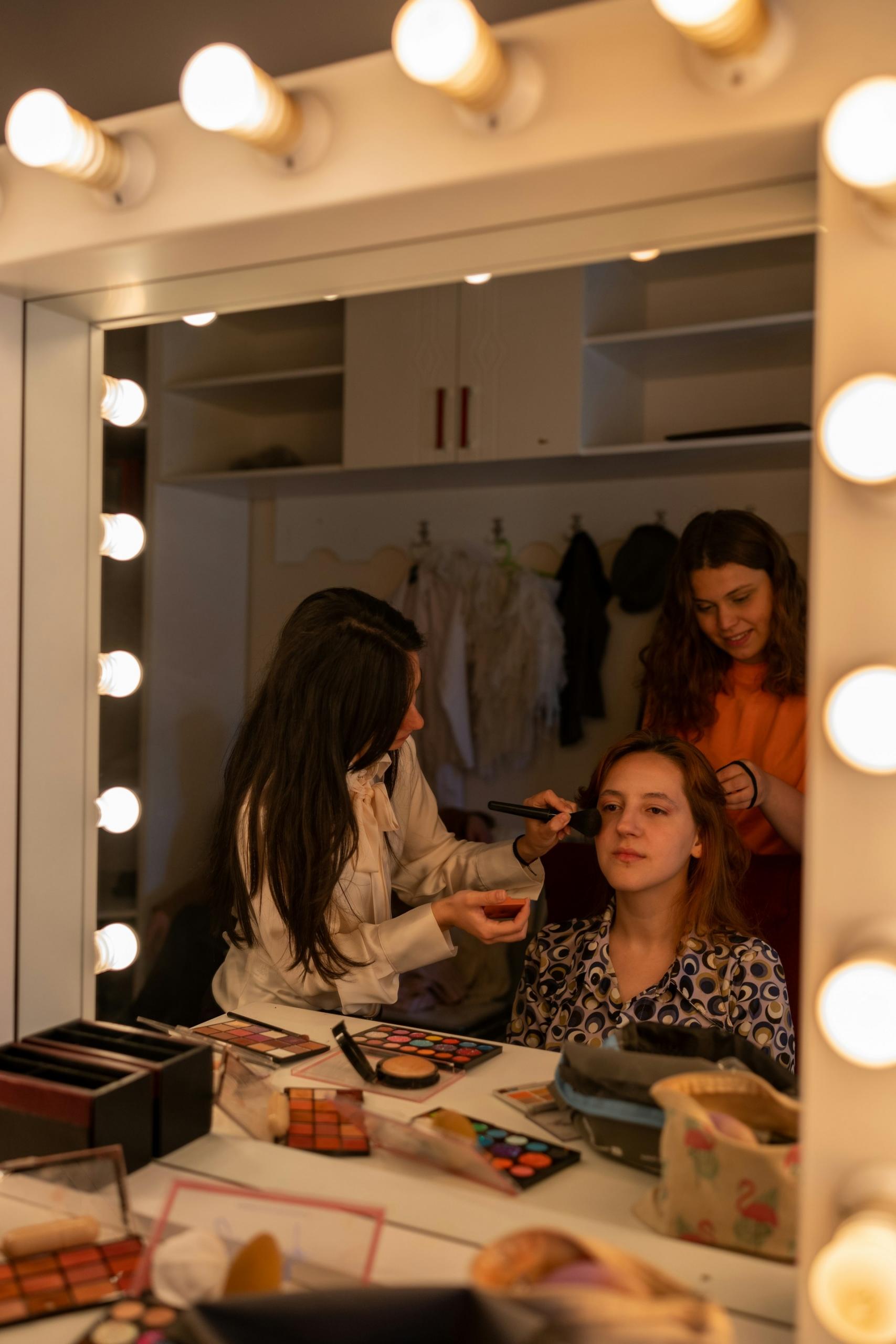 Two women fuss with another woman's makeup and hair in front of a mirror.