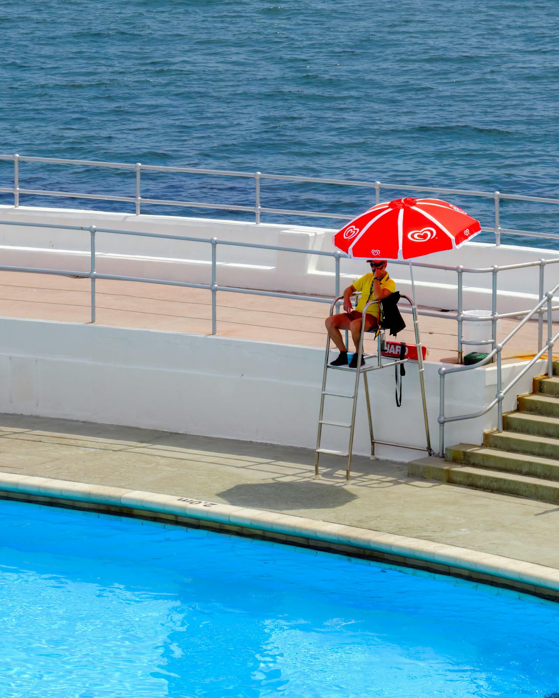 A man sitting under a red umbrella next to a swimming pool.