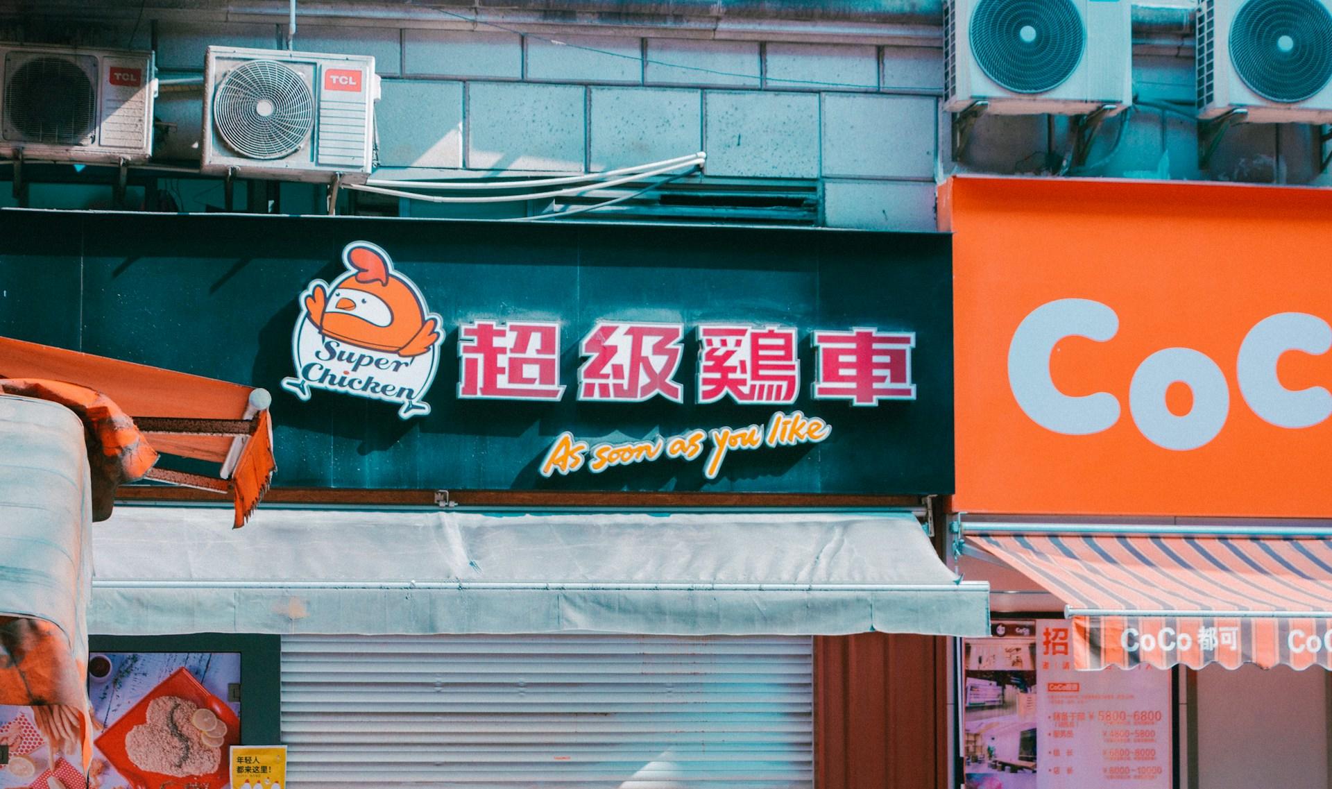 A close-up of a chicken snack shop sign featuring Chinese characters in red letters against a dark background, next to an orange cartoon chicken wearing a white apron.