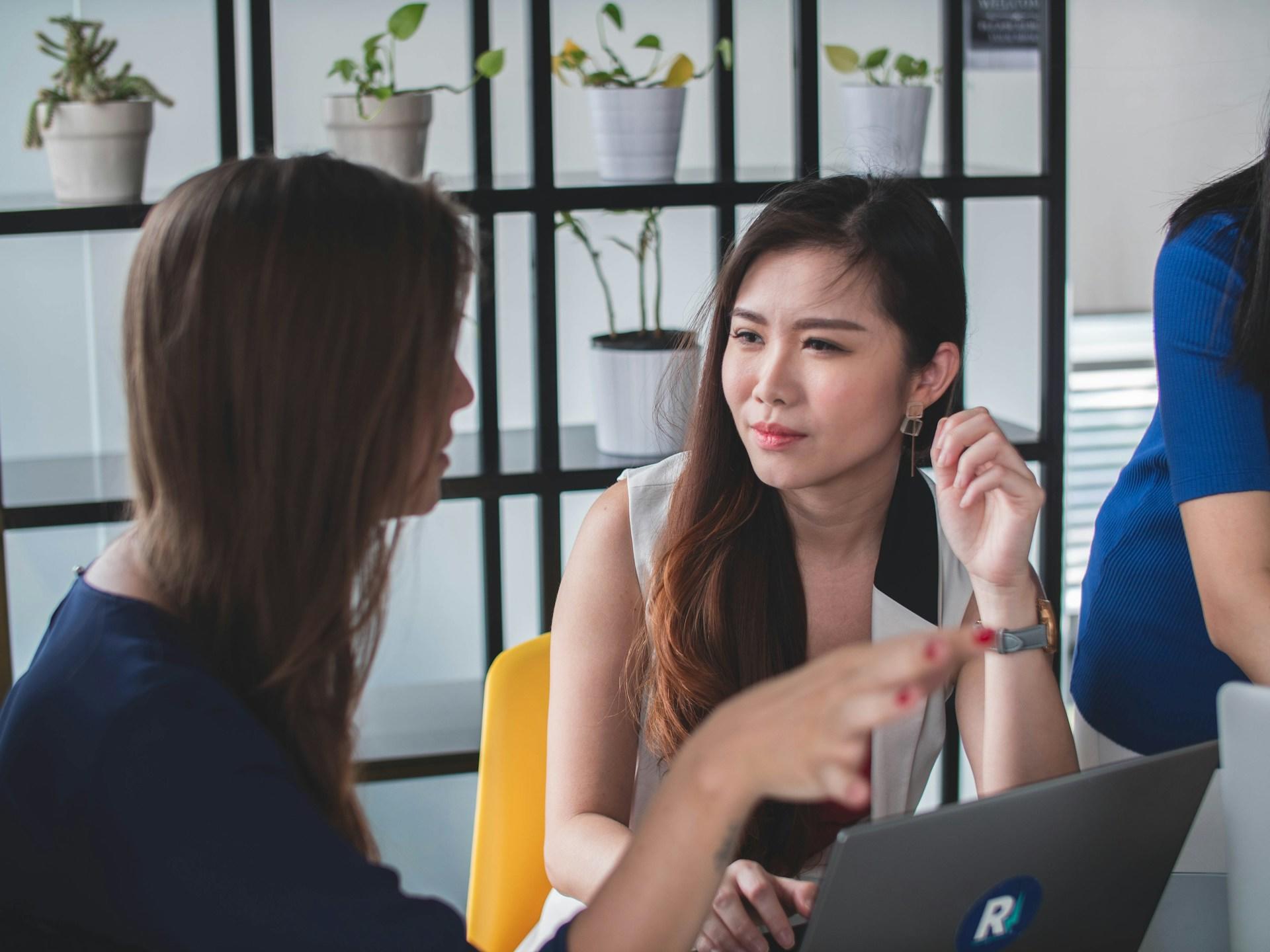 A person wearing a dark blue top sits next to a person with Asian features who appears to listen to the first person intently, ignoring the open silver laptop computer in front of them.