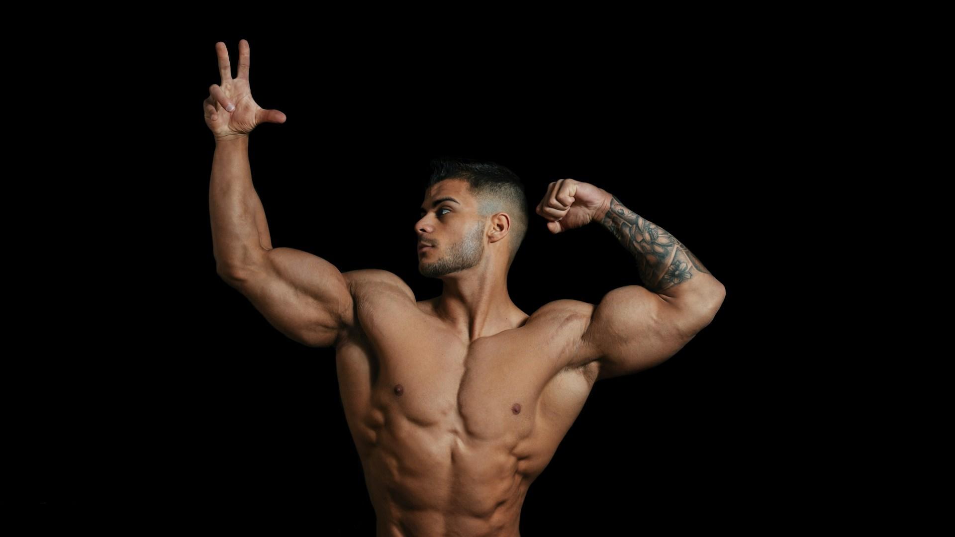 An extremely muscular mal displays his physique by striking a dramatic pose, shirtless against a black background.