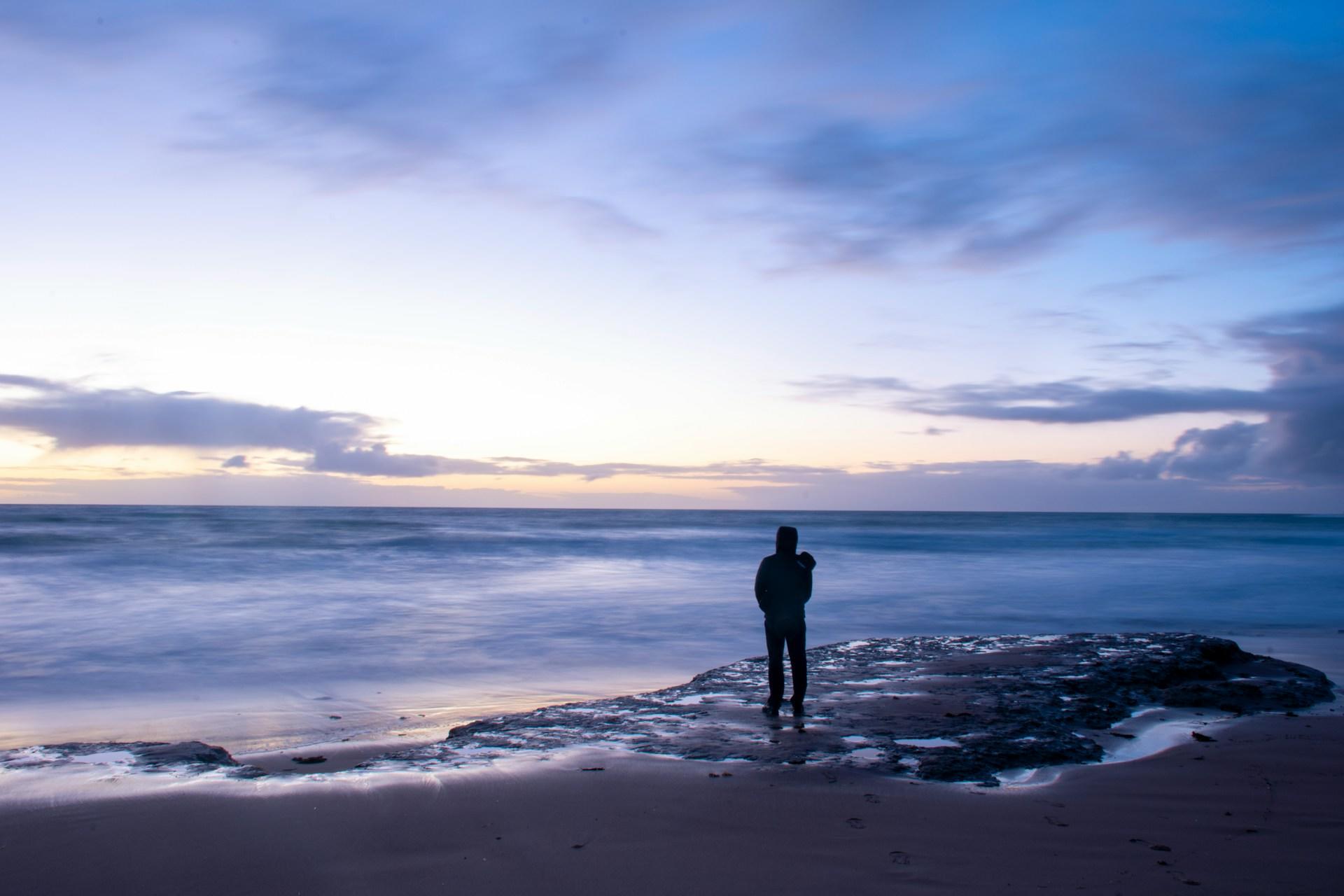 A person seen in silhouette stands on a rocky outcropping as water laps around their feet under a purple sky at sunset.