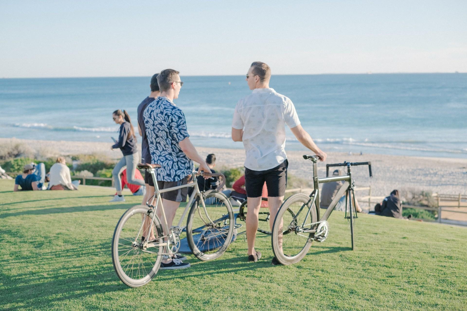Three people wearing summer button-down shirts and short pants stand next to their bikes on a grassy low bluff overlooking Scarborough Beach as other beachgoers sit or walk across the space.  
