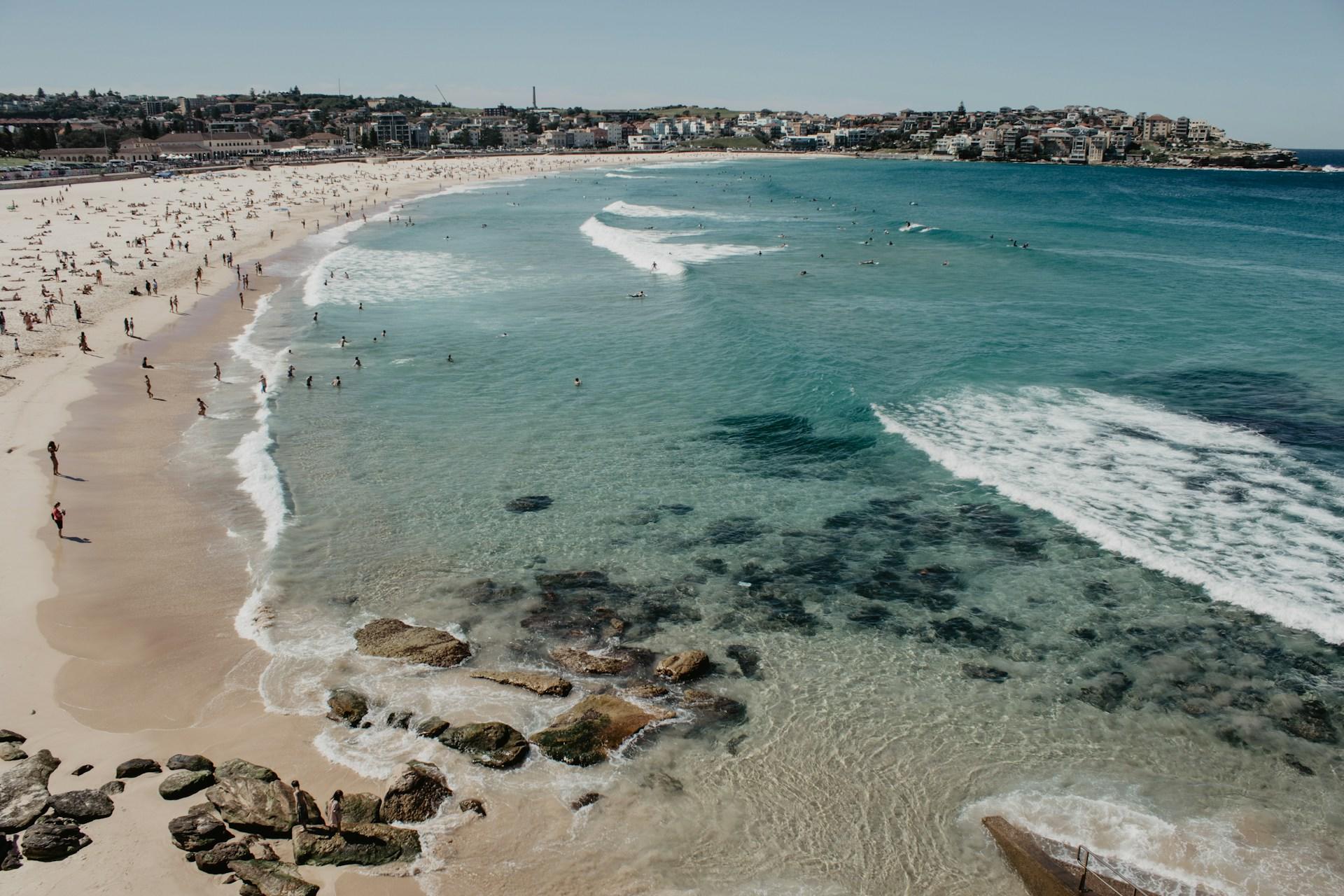 An aerial view of Bondi Beach in Sydney, showing turquoise waters and wavelets breaking on the sands, with rocks strewn in the foreground and people trolling along the tideline in the background.