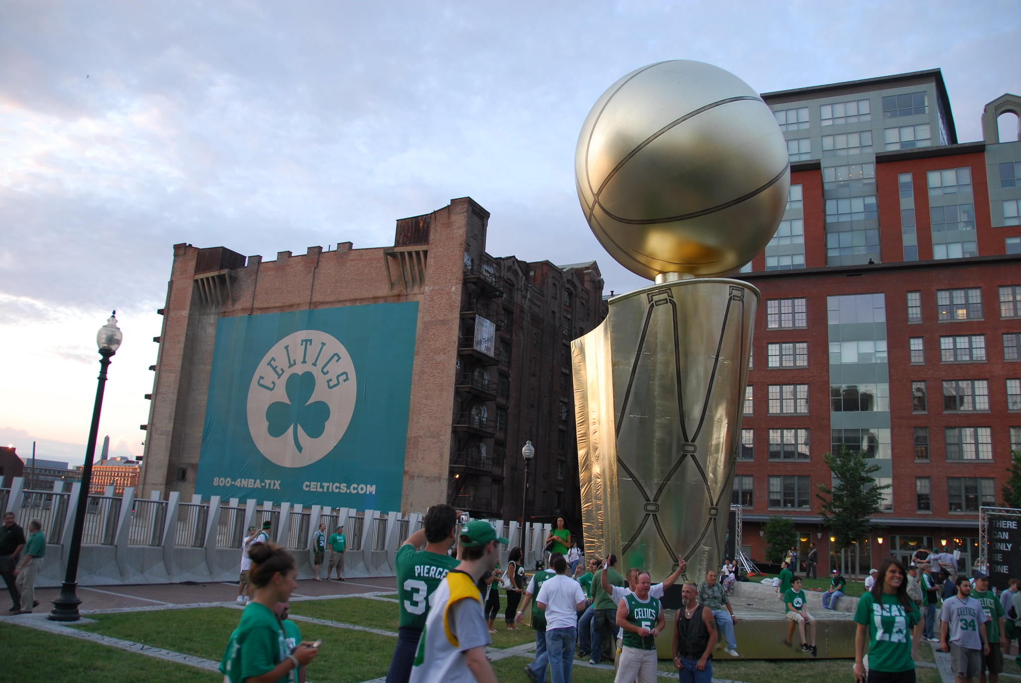 A giant temporary statue replica of the NBA championship trophy outdoors, in front of a giant banner advertising NBA tickets to the Celtics game. Many fans wearing Celtics jerseys are taking photos with the statue.
