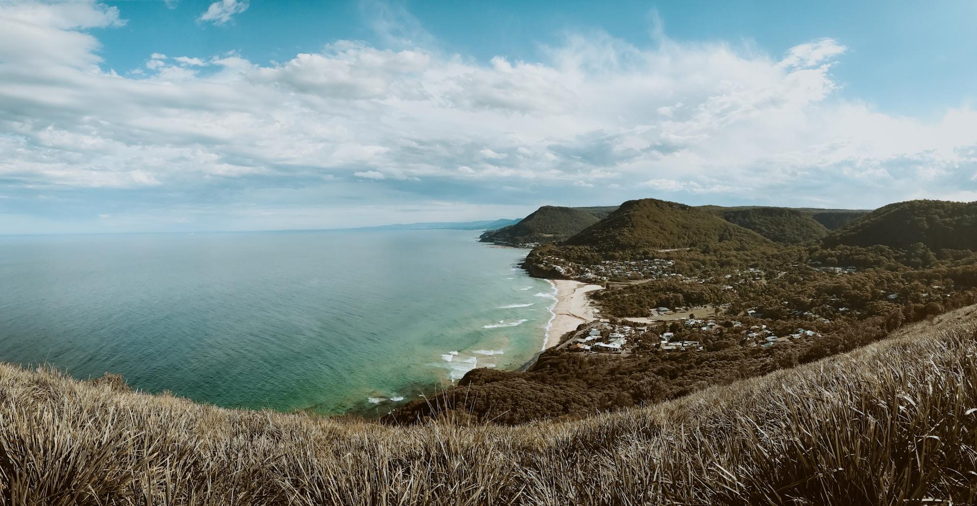 A view of Royal National Park beach seen from one of its peaks, with he waves making gentle forays onto the white sand and the scrub in the foreground looking brown and silver.