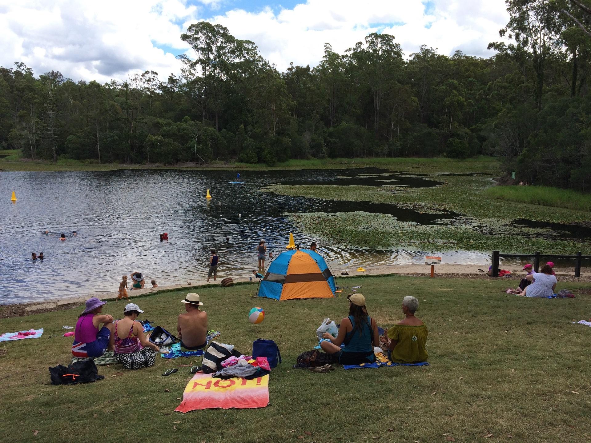 A scattering of people sit on the grassy banks of a body of water while some play in the water under mildly cloudy skies.