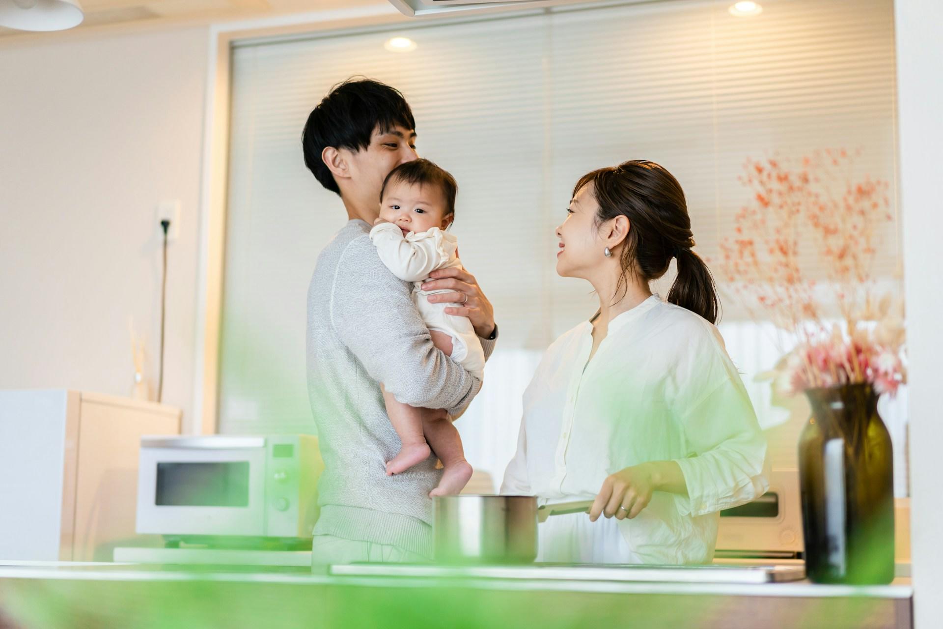 A man holding a baby and a woman stand in a brightly-lit space. They wear light coloured clothing and the woman holds a pot handle in her left hand while looking at the man and smiling.  
