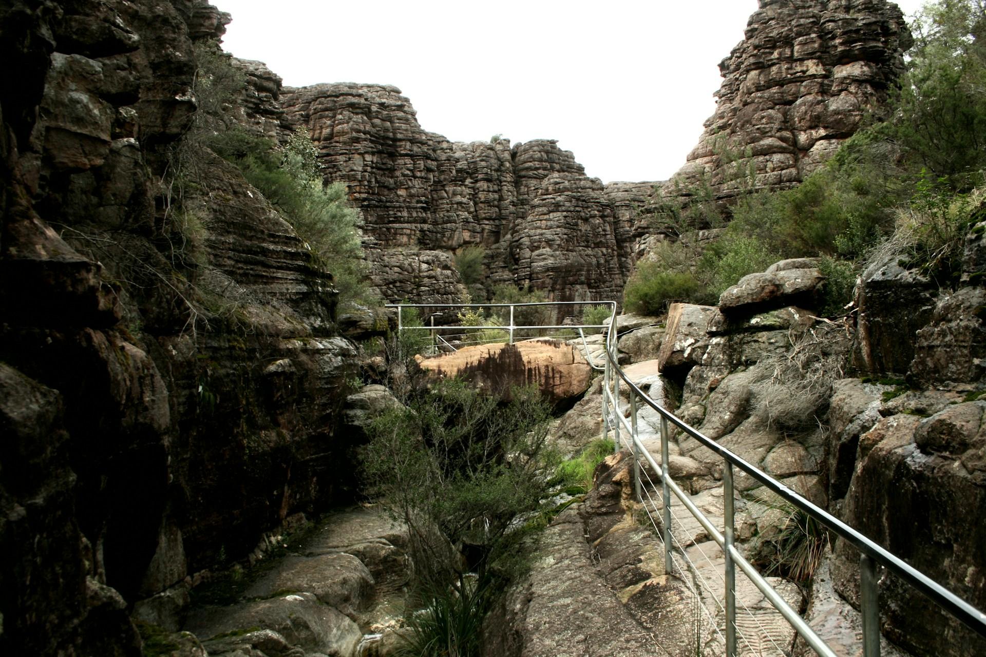 A narrow walkway cut into the rock face in Grampians Park, with its drop-off marked with a silver metal guardrail overlooking a rill of water below it.   