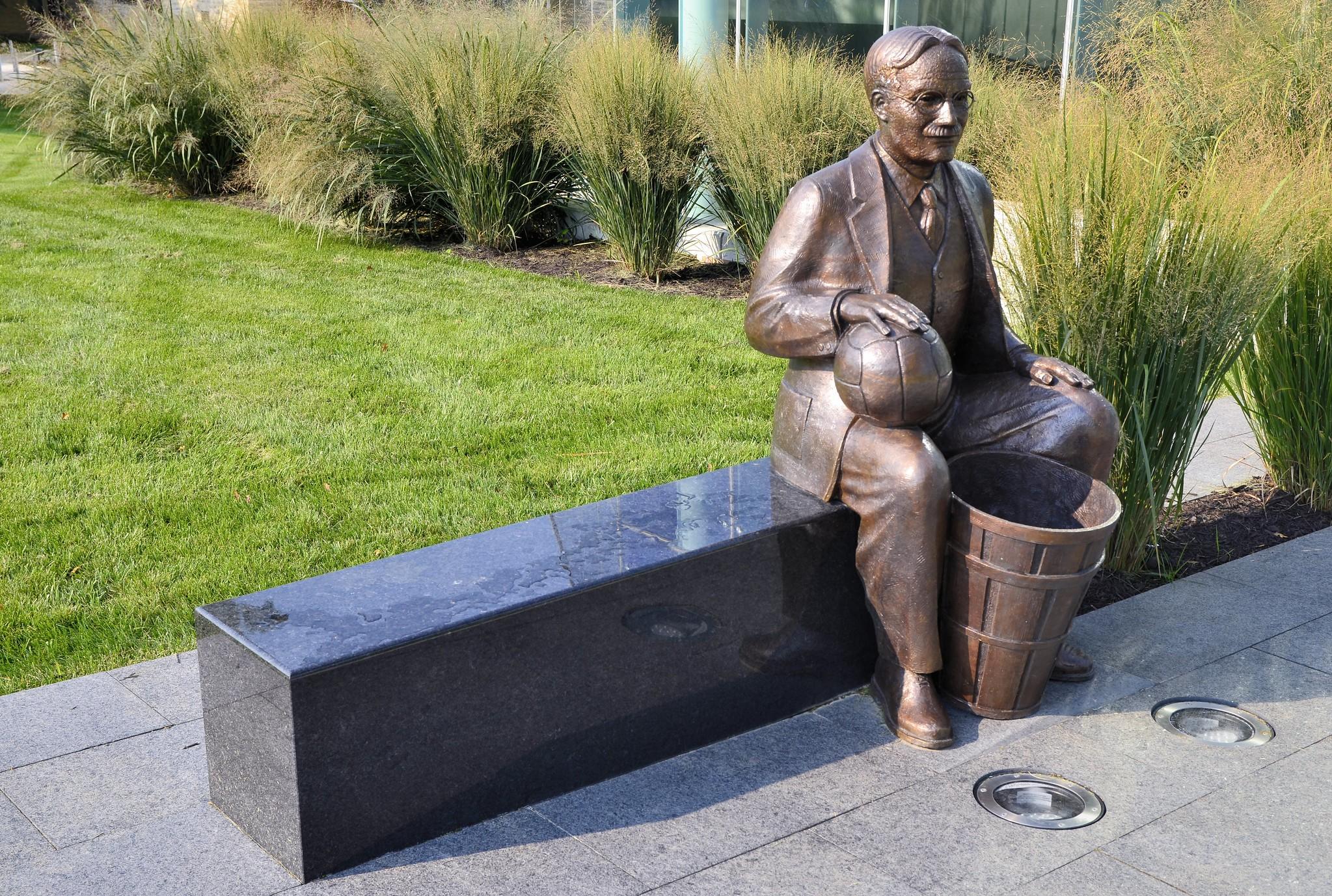 A bronze sculpture of James Naismith sitting on a bench and holding the first iteration of a basketball and peach baskets.