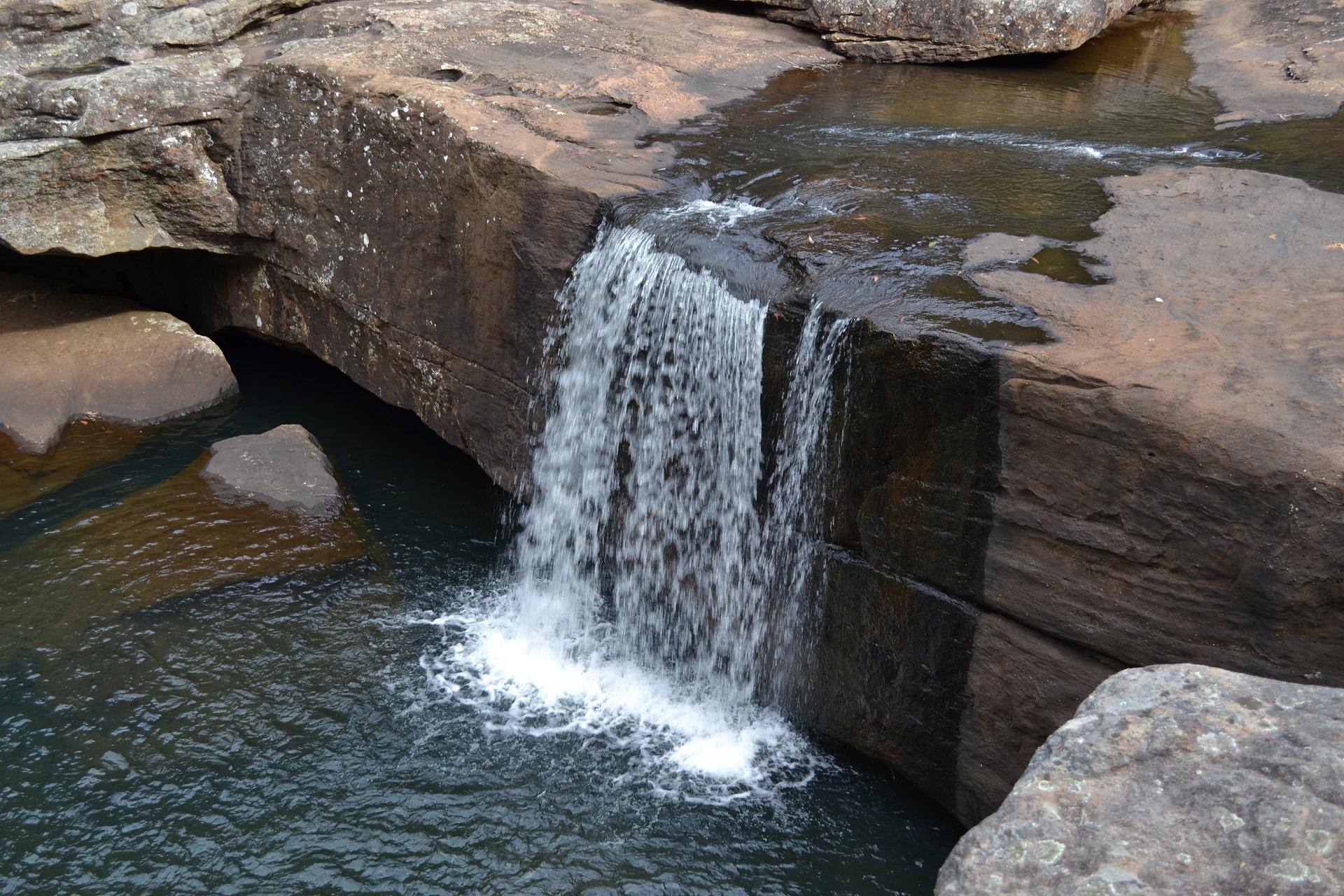 A small cascade of water flows into a green swimming basin from a large flat chunk of reddish sandstone.