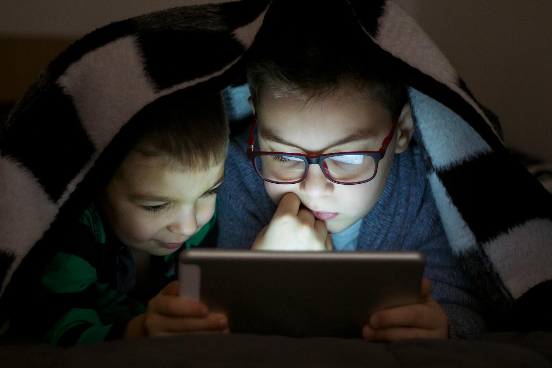 Two small children, one wearing black and red framed glasses, huddle under a black and white checked blanket, their faces lit up from a tablet they're focused on.