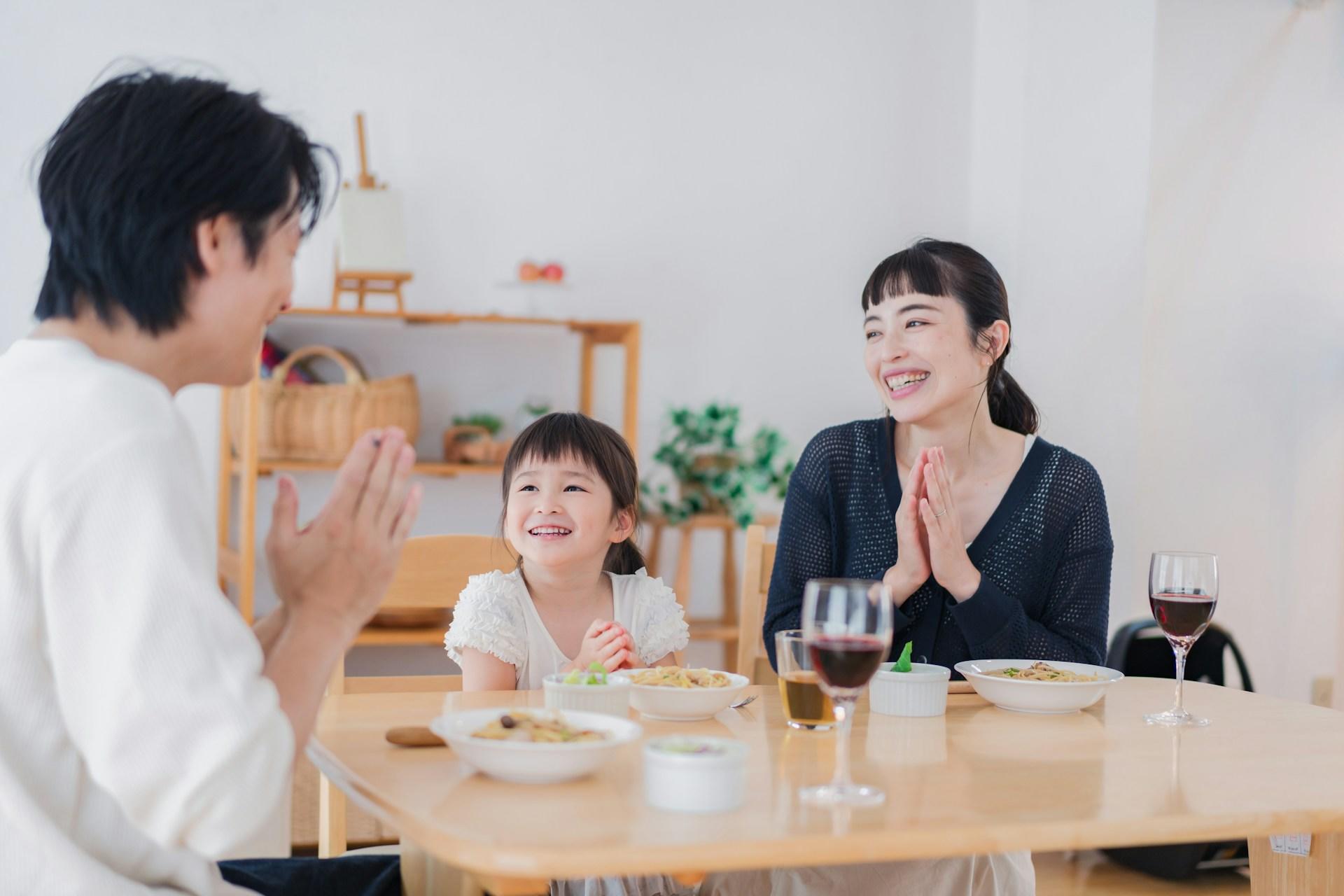 A man wearing a white shirt sits at a blond wood table with a woman wearing a dark top and a child wearing white. The table holds wine glasses and other dinnerware. The family laughs and smiles.