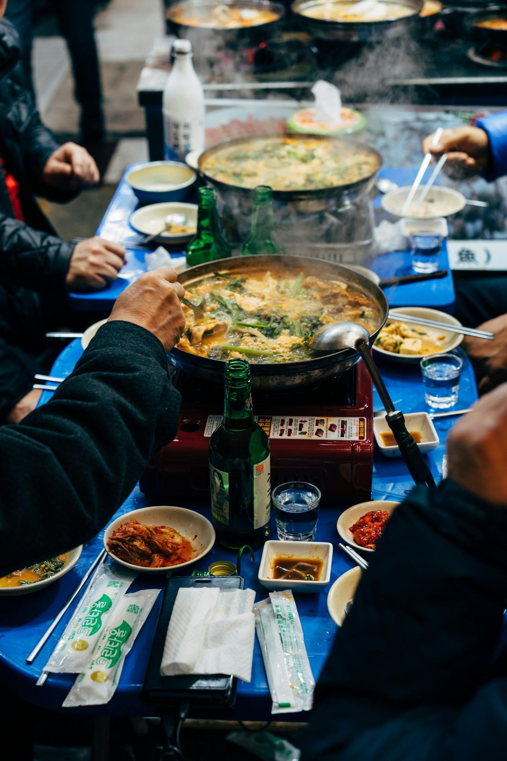 People sit around a blue-topped table laden with banchan and stew kept warm with a gas burner. Their winter clothing and the steam rising off the stew gives the idea that it must be cold. 