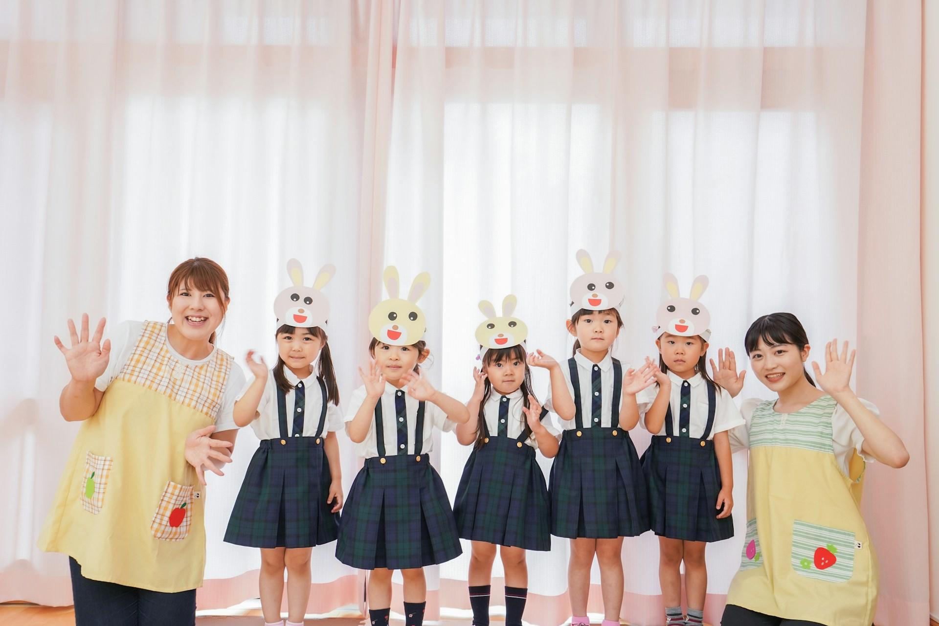 Five small schoolchildren wearing bunny masks about their faces stand in a row between two teachers wearing yellow pinafores in a brightly lit room.