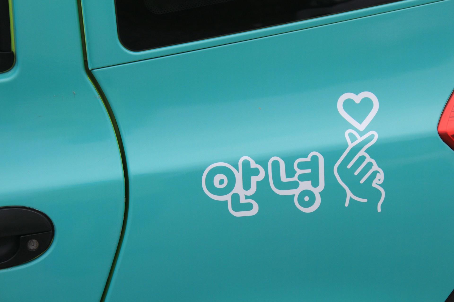 A teal coloured car's quarterpanel displaying a white sticker in the form of the Korean word for 'hi', next to a hand making a heart gesture and a heart above the crossed thumb and finger.  