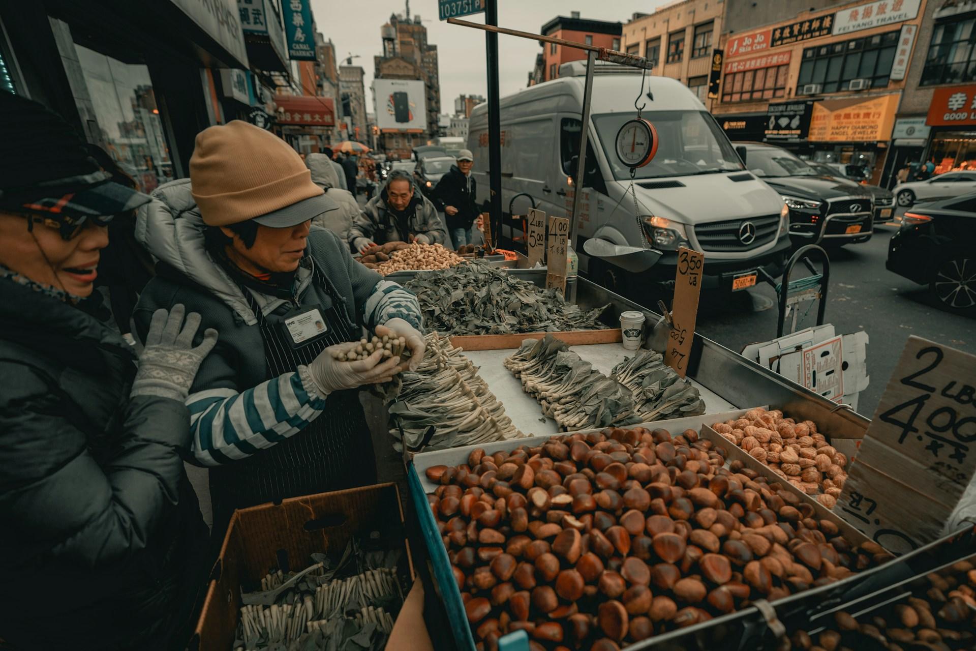 Two bundled up Korean elderly women stand behind an open-air stall, stocking chestnuts and various vegetables on a crowded street on a gloomy day. 