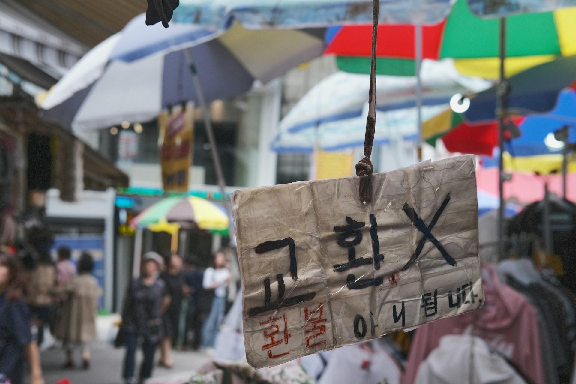 A handmade sign with Korean writing in black and red ink hangs by a thin strap in front of an open market stall.