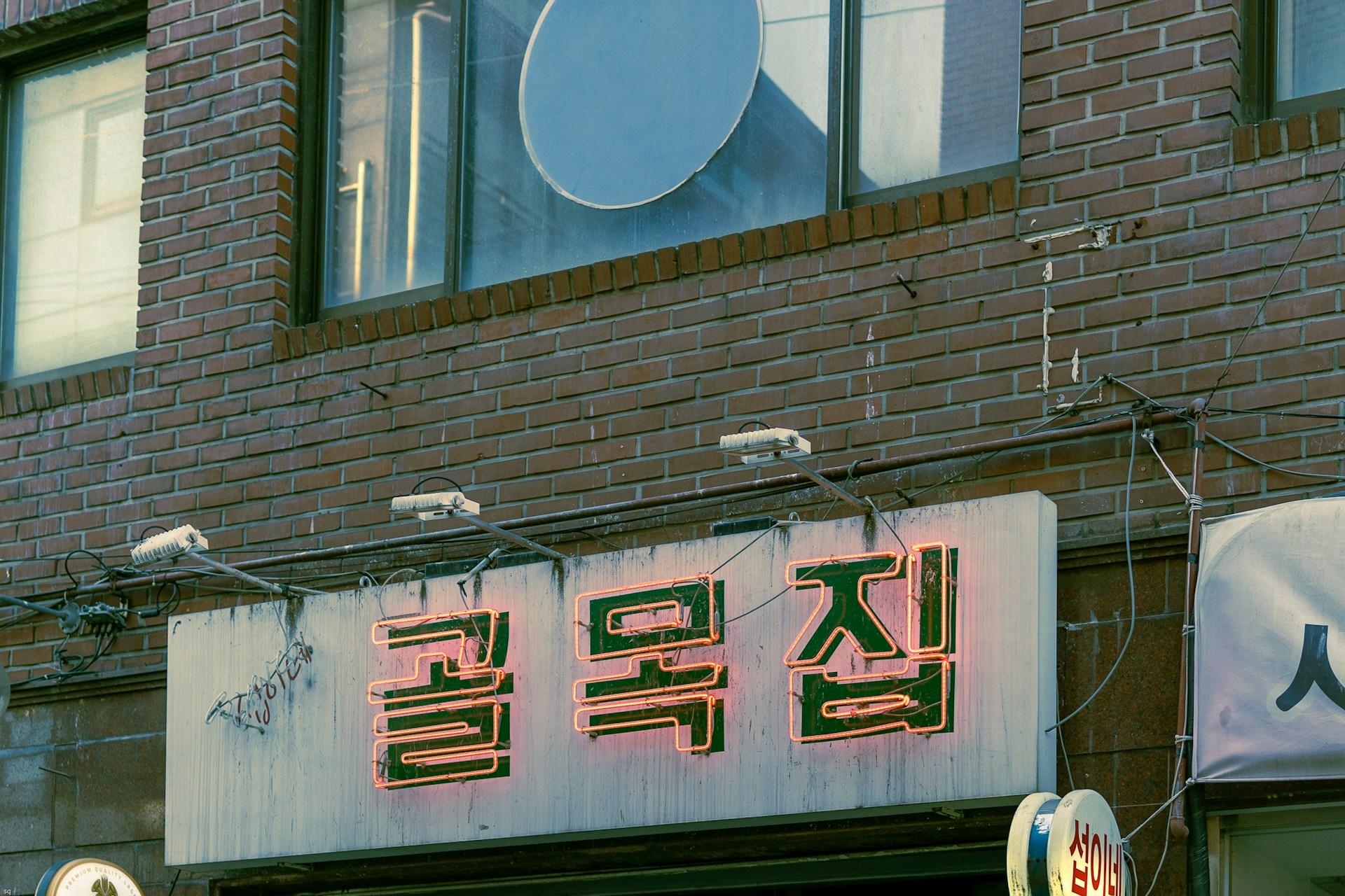 A building with a brick facade featuring a white sign with green Korean writing surrounding by golden neon.