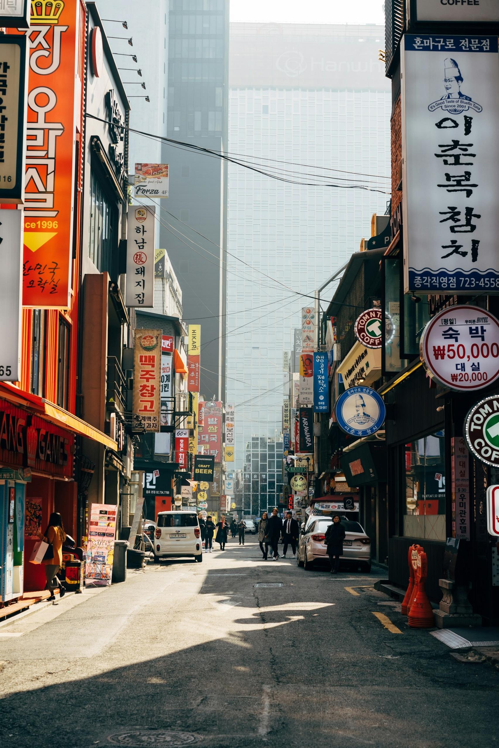 A narrow street in Seoul with colourful vertical signs advertising the shop and services they hang over, with a few pedestrians in the distance and a glass-faced building in the background.  