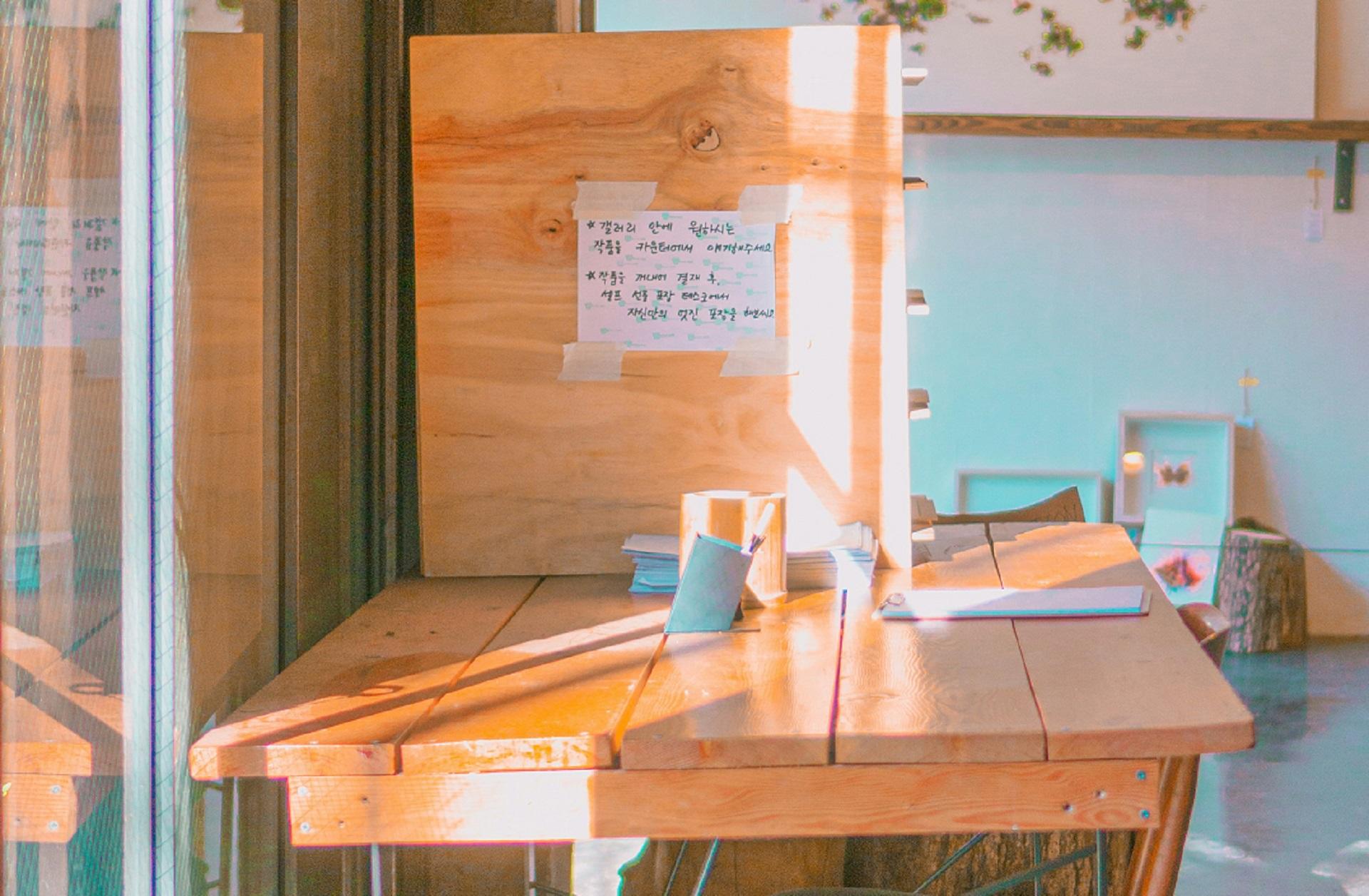 A wooden desk with an upright partition abutting a glass door, through which sunshine pours in. On the partition a paper with Korean characters is taped.