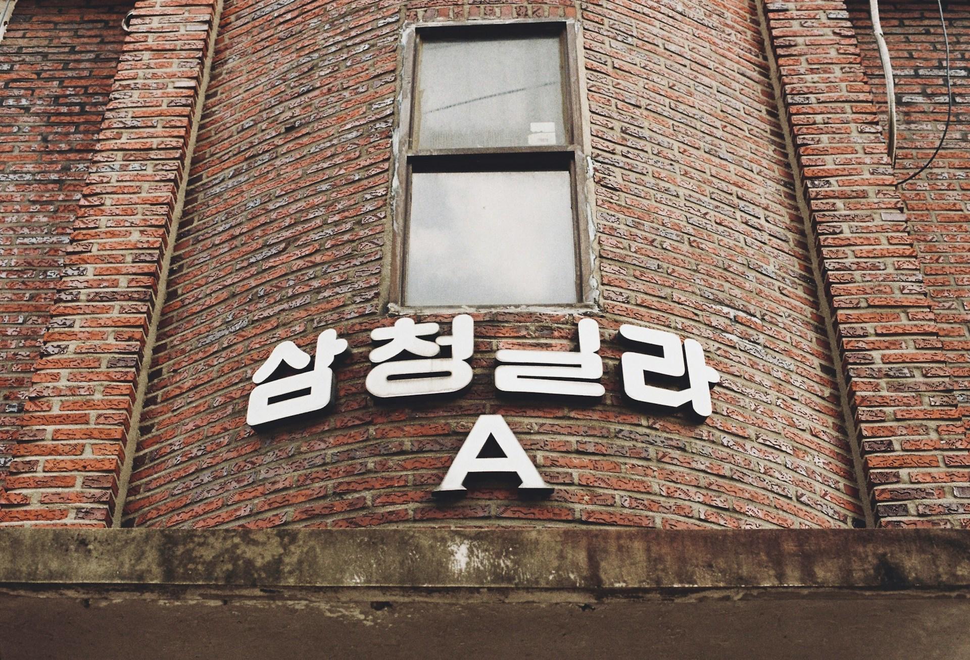 A brick building facade with a convex feature boasting a window and several Hangul, plus the letter A, in white. 