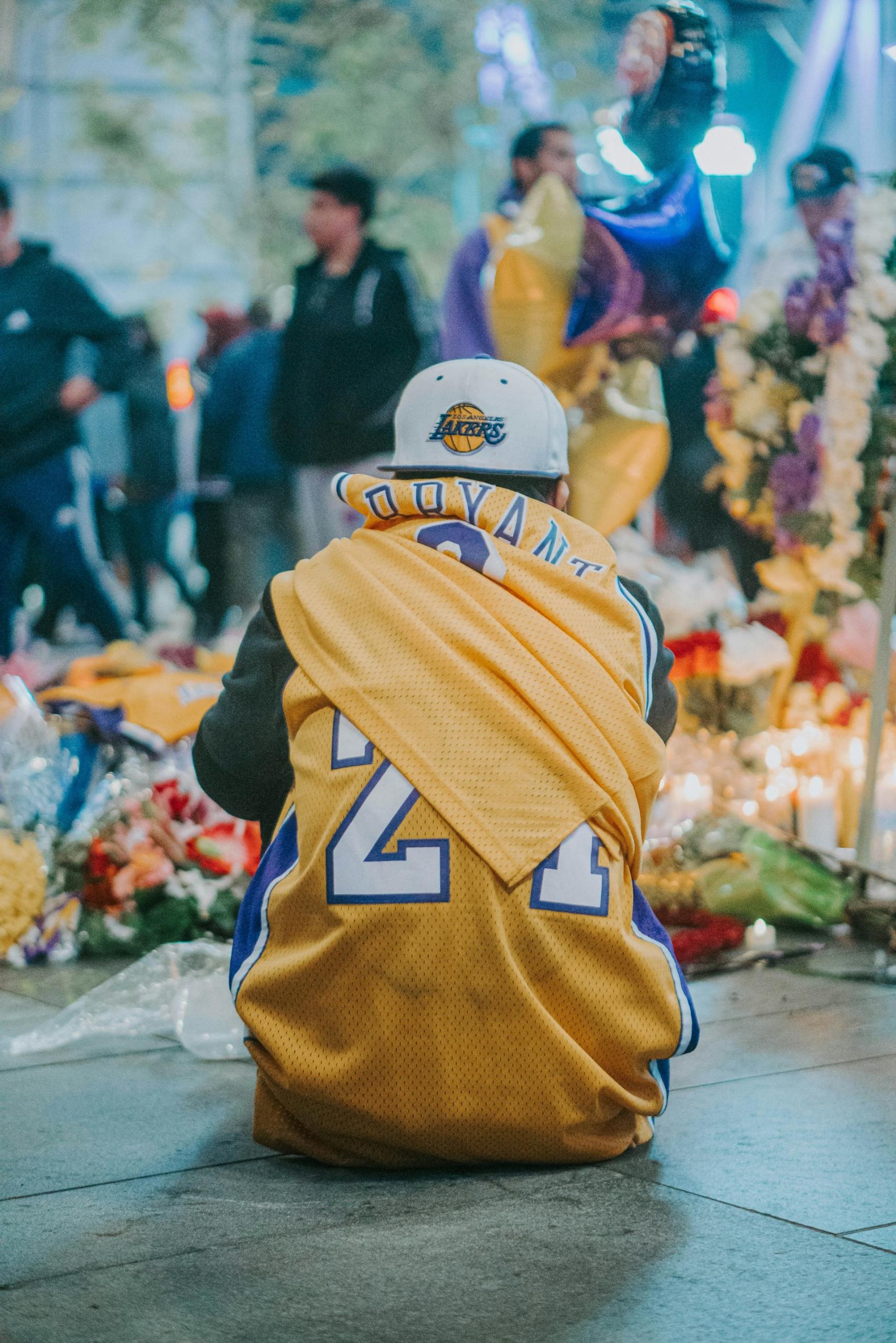 A photo of a fan wearing an LA Lakers jersey sitting on the ground in front of many flowers laid down by fans in tribute of Kobe Bryant.