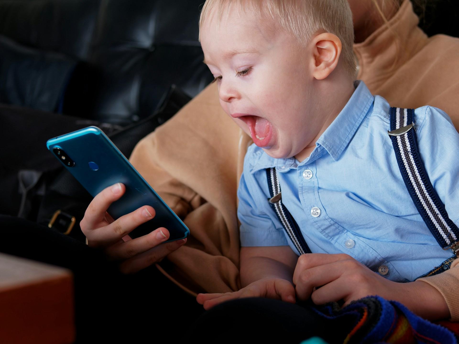 A blond toddler wearing a light blue button down shirt with blue, white and grey suspenders sits on a person's lap, watching the blue-cased smartphone the adult holds, with their mouth wide open in amazement.