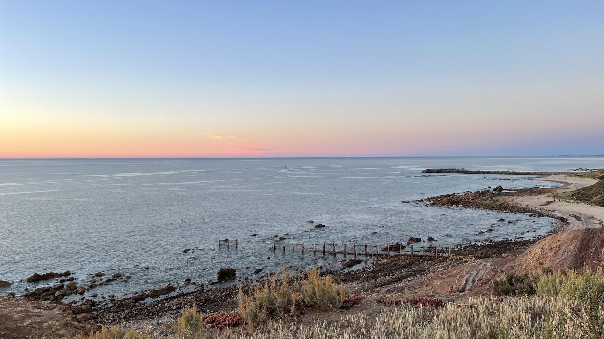 O'Sullivan Beach in Adelaide, seen at sunrise from atop a bluff, showing its pier and rocky outcrops stretching into the water. 