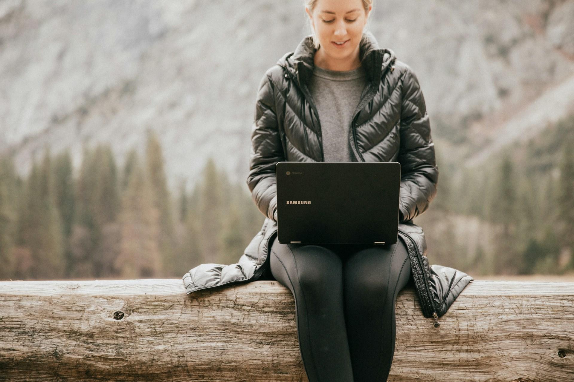 A girl sitting on a log in the wilderness using her laptop