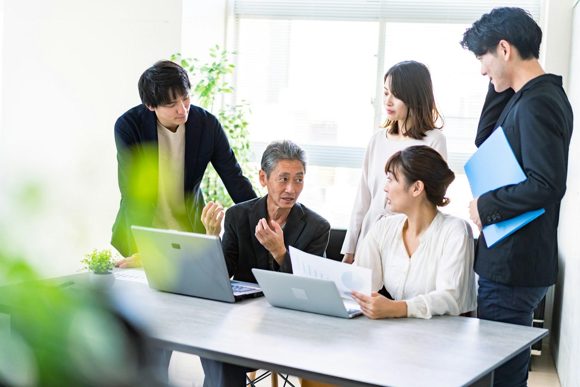And elderly man sits at a grey table with an open laptop computer in front of him. Four younger people stand around him, focused on him as he explains something. 